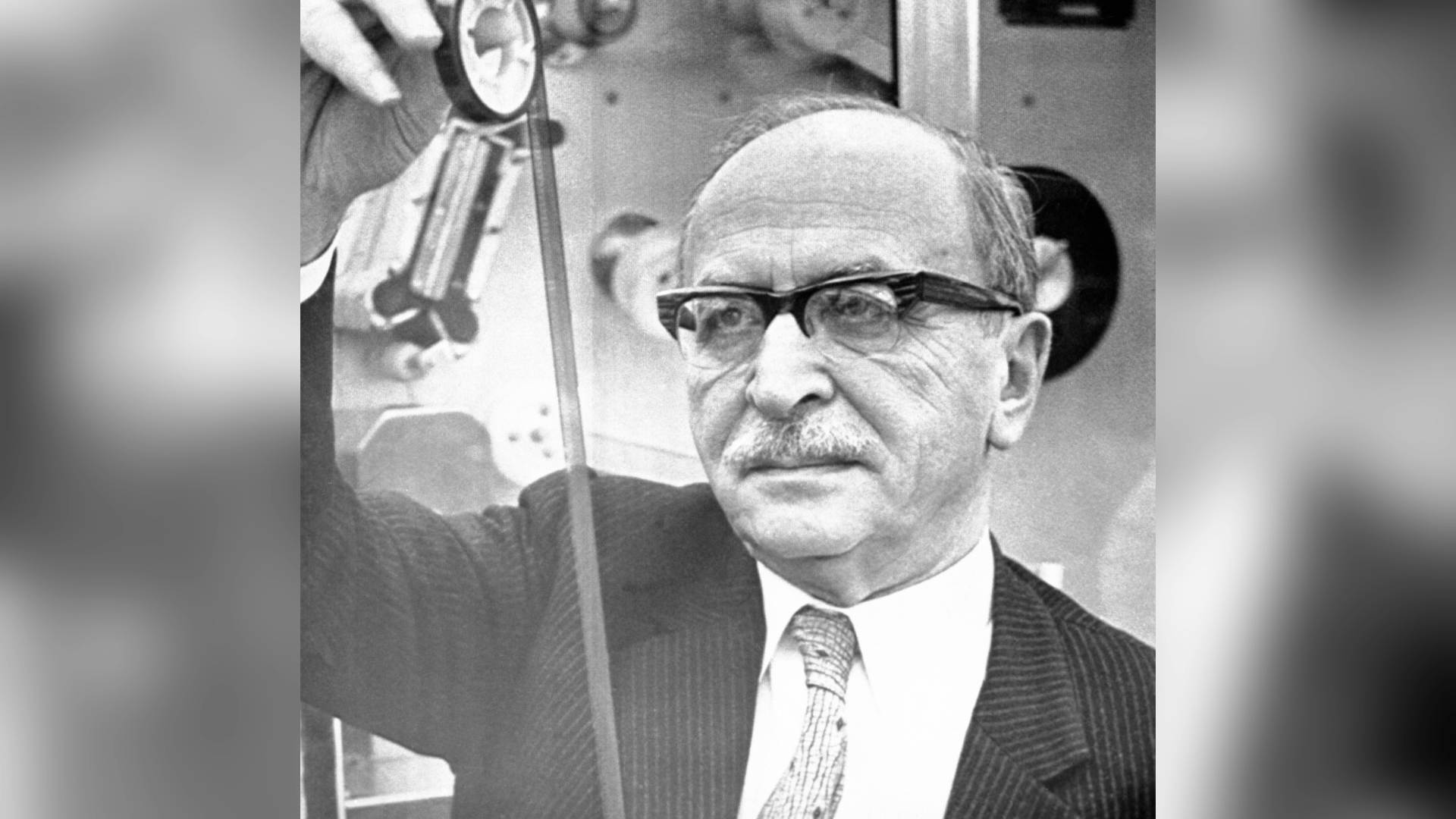 A black-and-white image of a man wearing glasses. He is holding up a measuring device and appears to be in a science laboratory or workshop.