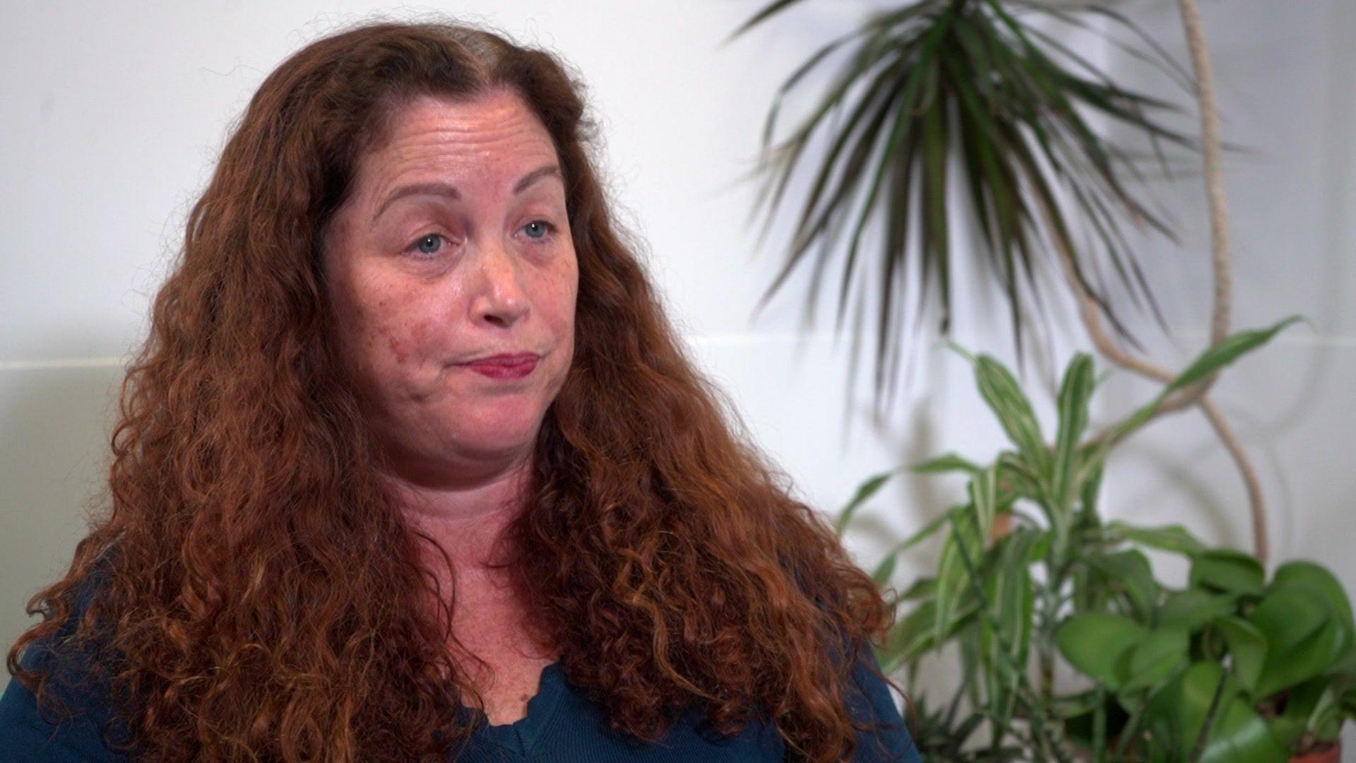 Dr Nicola Connolly is sat down wearing a blue top. She has long frizzy ginger hair. Behind her is a neutral wall and some plants.