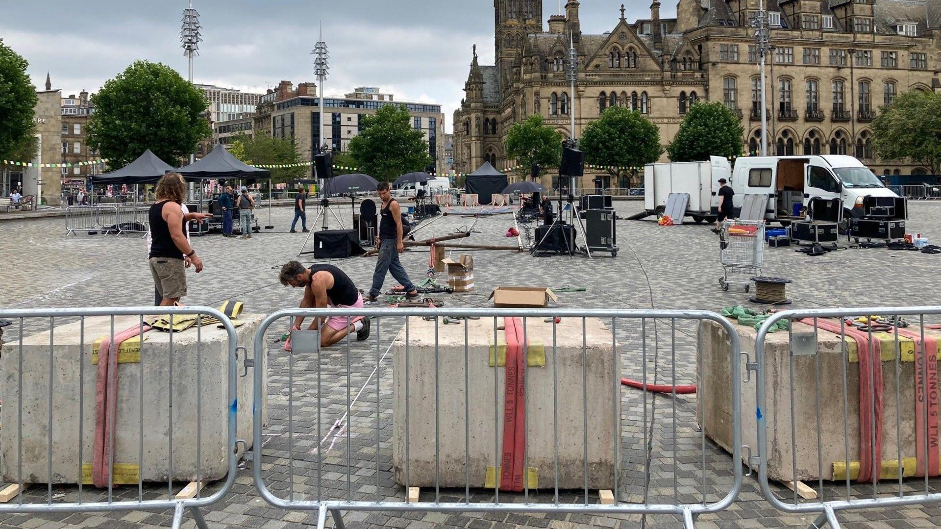 Metal railings in the foreground with people setting up the high wire performance space with the Bradford City Hall clocktower in the background.