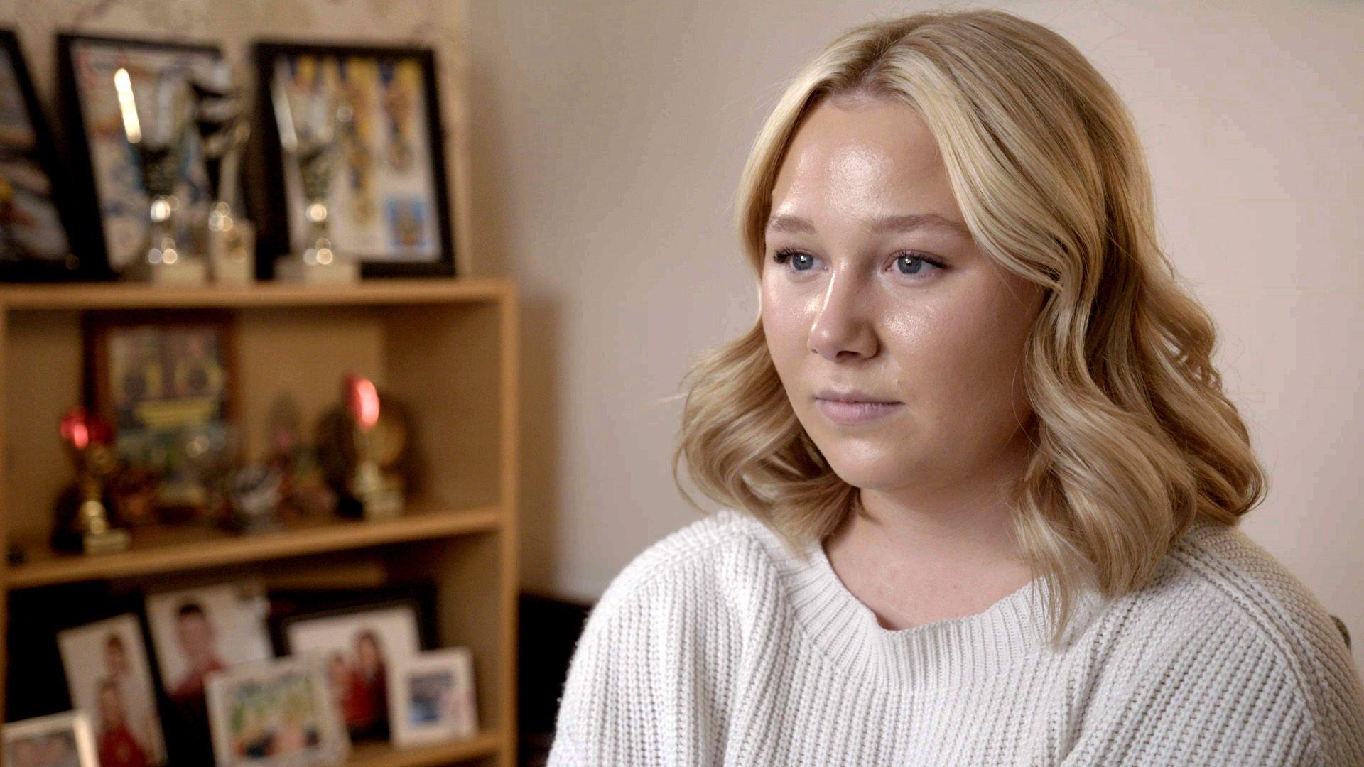Headshot of Abby, who is pictured looking at a camera to the left hand side of the shot. She has shoulder-length, blonde, wavy hair and is wearing a cream ribbed jumper. She is pictured next to a wooden bookcase containing framed photographs and trophies.