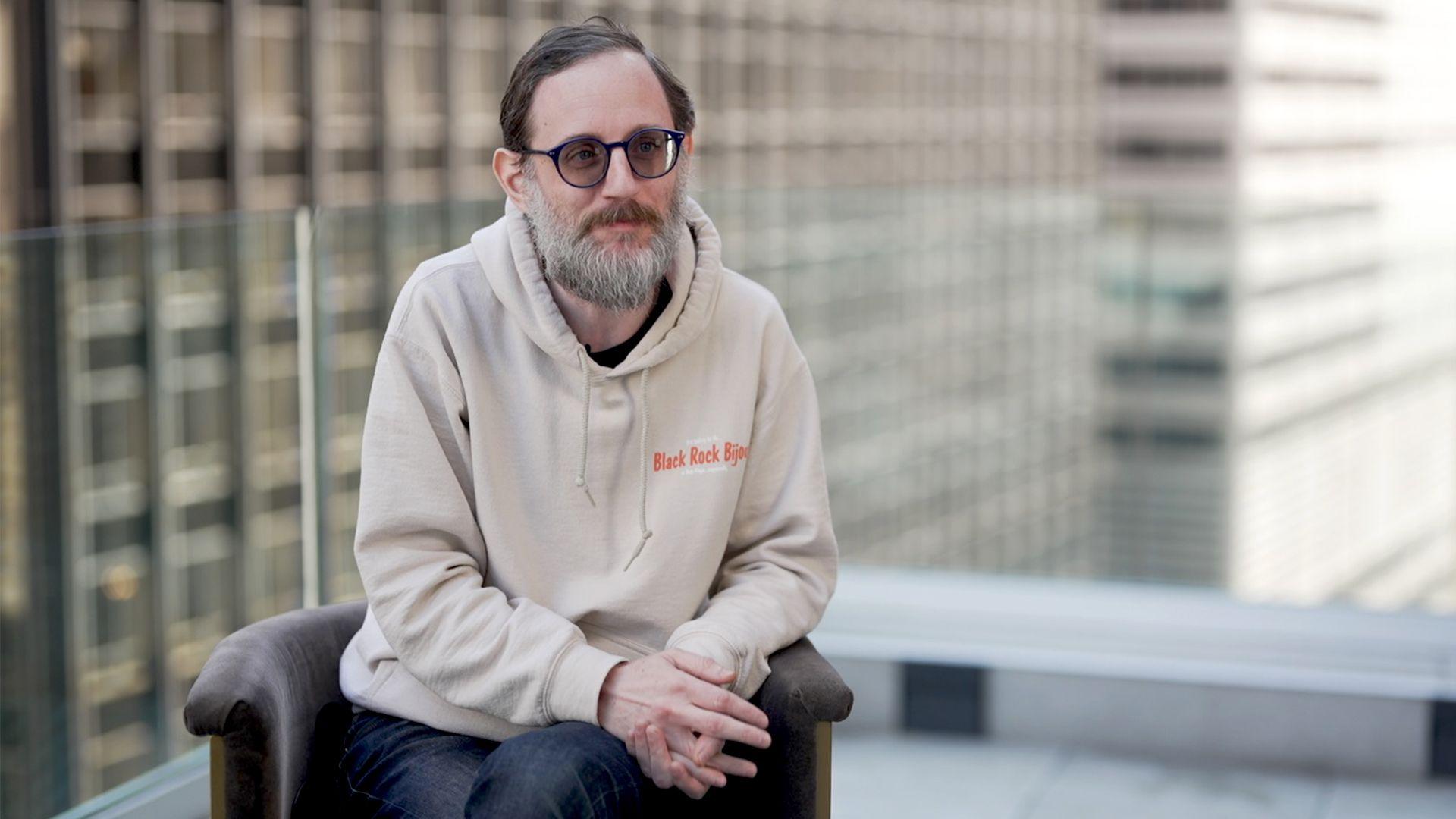 Luzer Twersky leans forward, hands clasped, dressed in a beige hoodie