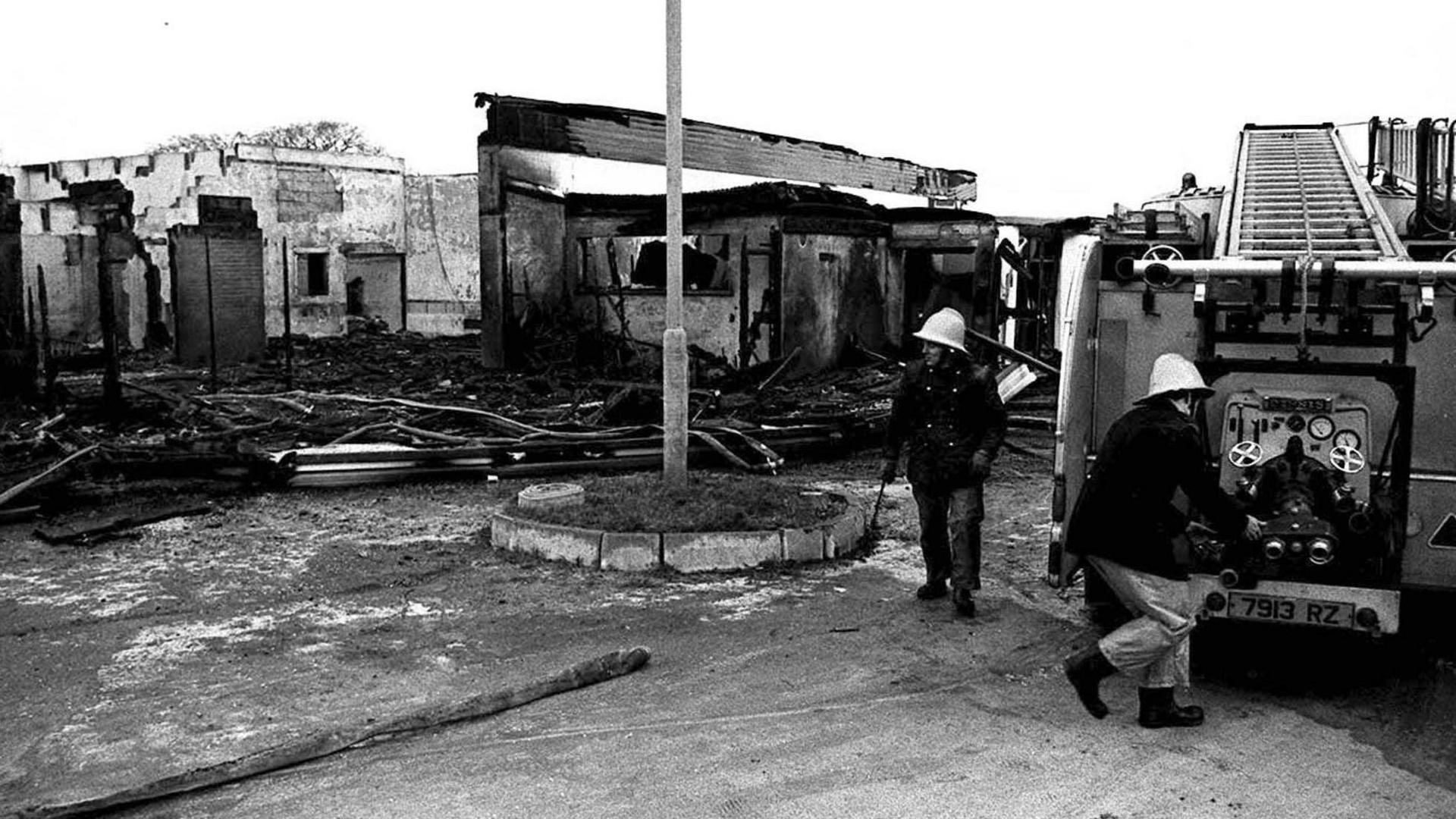 Black and white image of the bombed La Mon hotel, the brick building is covered in black with parts with most of the building frame collapsed, debris lies on the floor and two men stand by an old fire engine outside the building they are wearing dark jackets and trousers with black wellington boots and  hard hats. One man is getting the hose from the back of the fire engine.