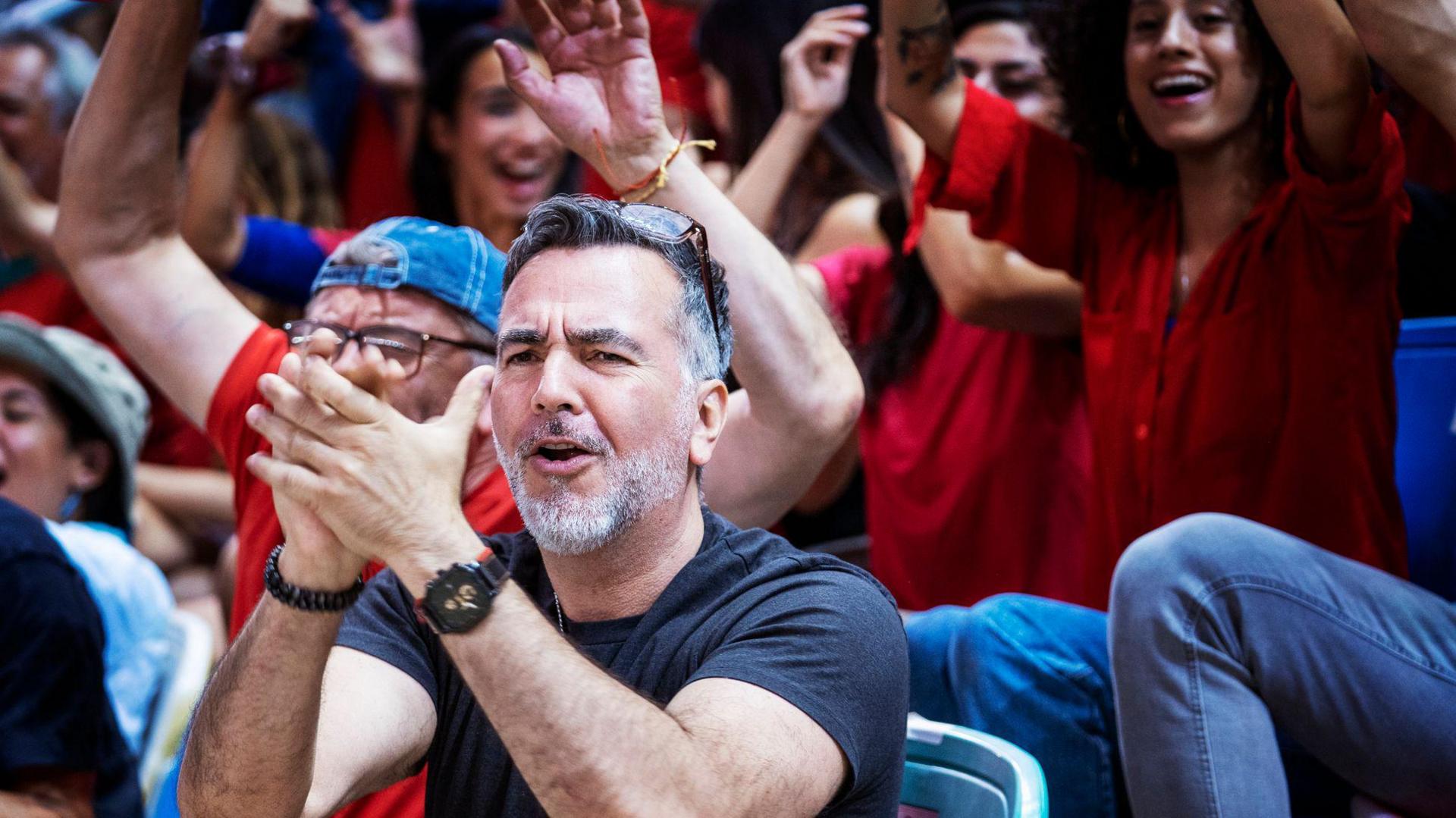 Enthusiastic audience of sports fans in support of the home team sit in stadium chairs and raise their arms while cheering and clapping