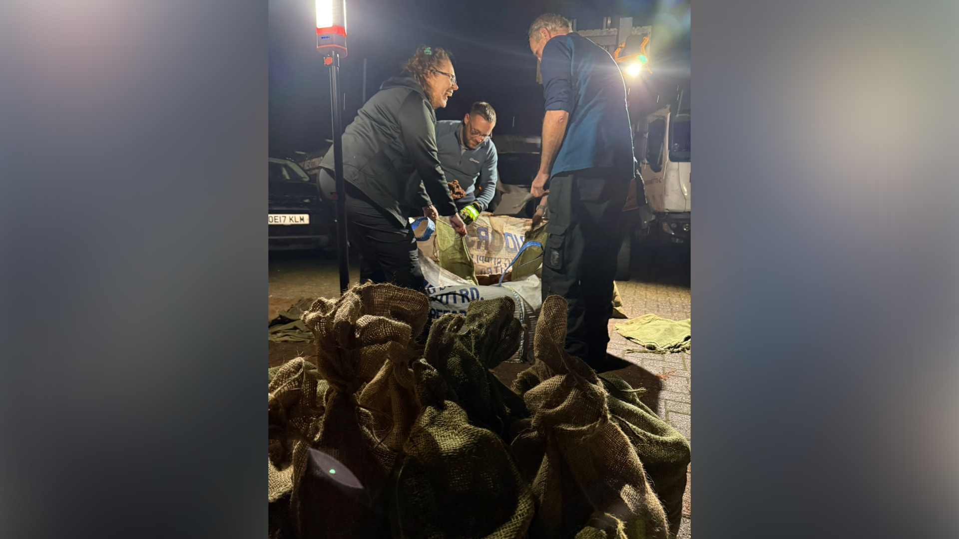 Three people pictured in the foreground away from a pile of sandbags. The image is taken at night with large spotlights lighting up the area.