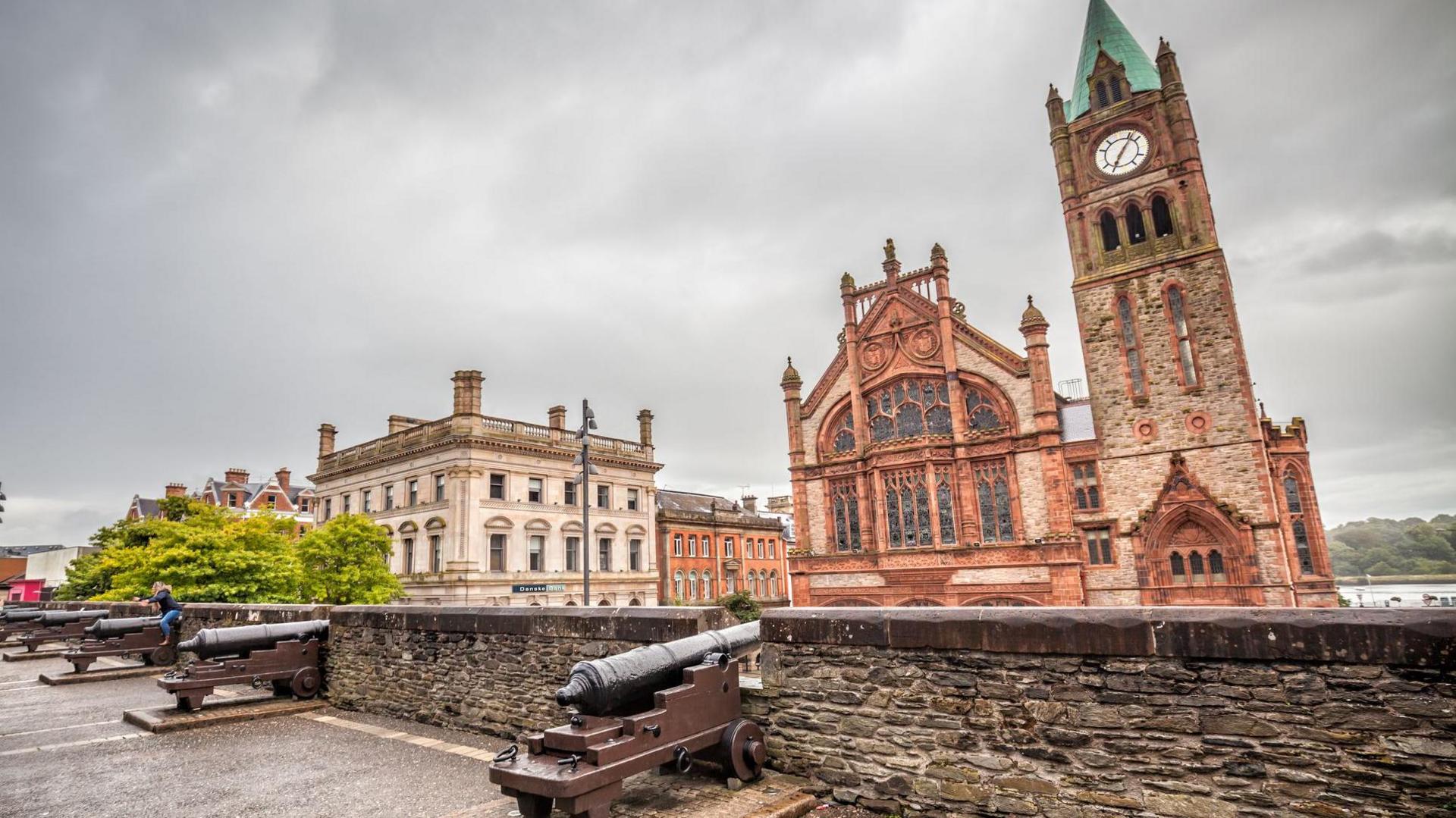 In the foreground on the wall we have several cannons sticking out between the battlements. A girl accompanied by a boy takes pictures of another girl who has climbed a cannon on the wall To the right outside the wall is The Guildhall, building located next to the river, very close to the center of the city, built in 1887 of sandstone and brick. 