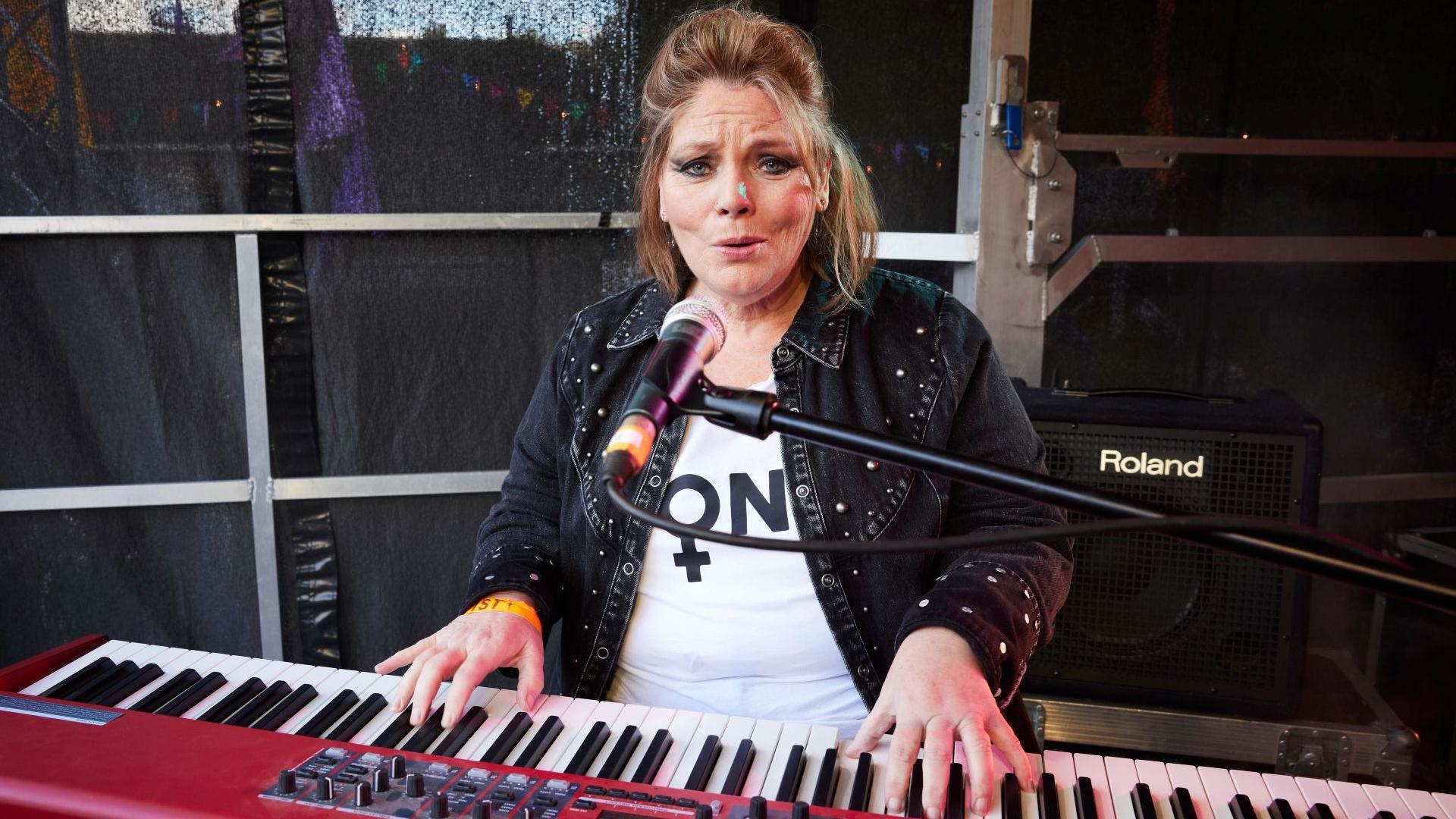 Joanna Scanlan sits at a red electric keyboard, with her hands above the keys ready to play. She is wearing a black denim jacket and white t-shirt. She has dark eye make up. Her lips are open as though she is singing.