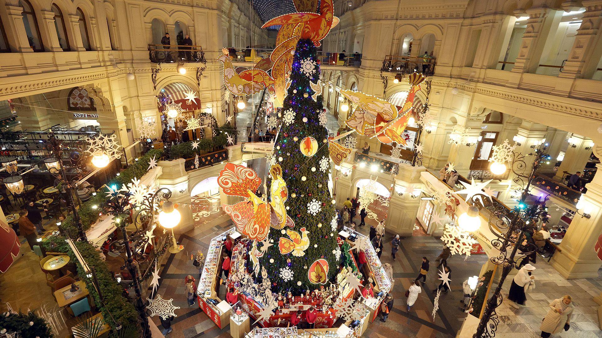 a christmas tree decorated with brightly coloured shapes stretches high in the foyer of a shopping mall with shoppers walking around it