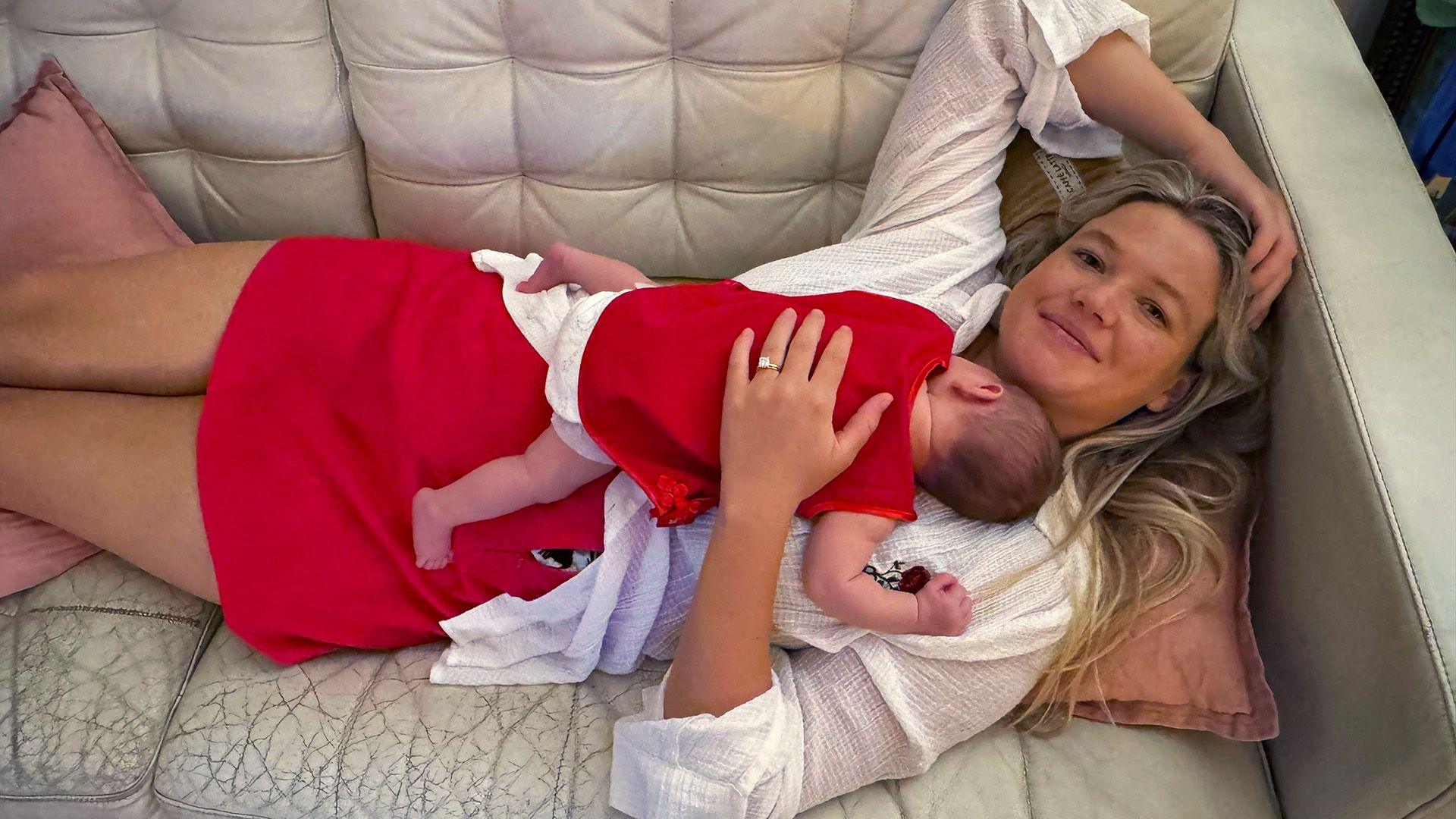 A woman with long blonde hair, wearing a white top and red skirt lying on a cream couch, with a baby sleeping on her chest.