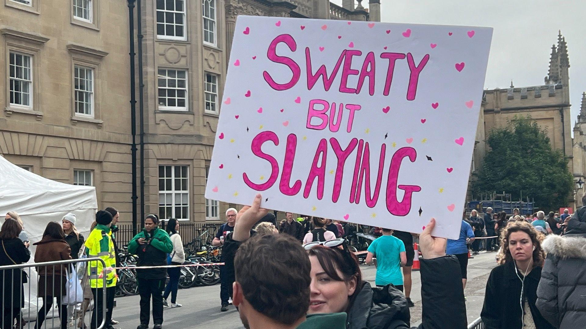 A woman holding a sign up that says 'sweaty but slaying' in pink and gold letters.