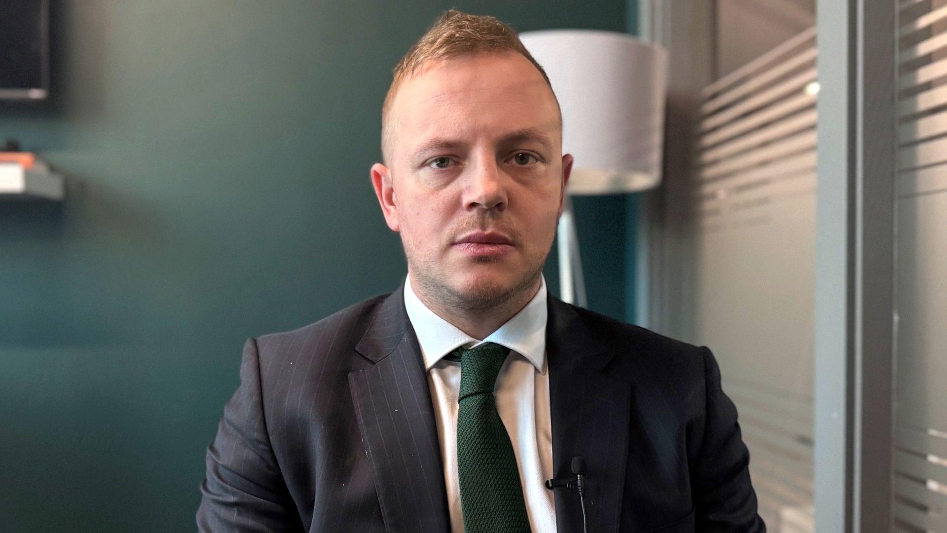 A man with short blond hair looks towards the camera. He is wearing a dark coloured suit jacket, a white shirt and a dark green tie. He is sitting in an office with a dark green wall in the background.