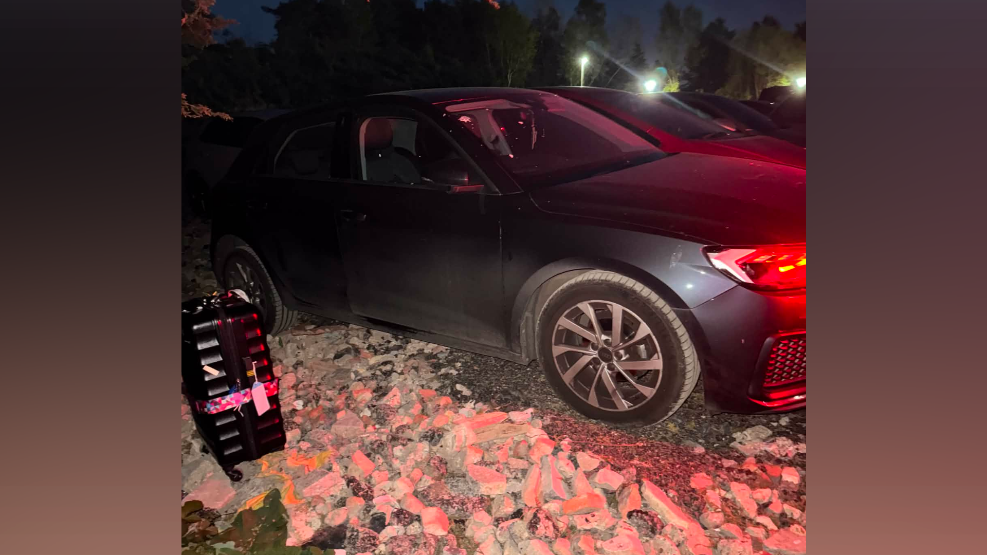 Black car parked on gravel and broken concrete at night with a suitcase next to it. 