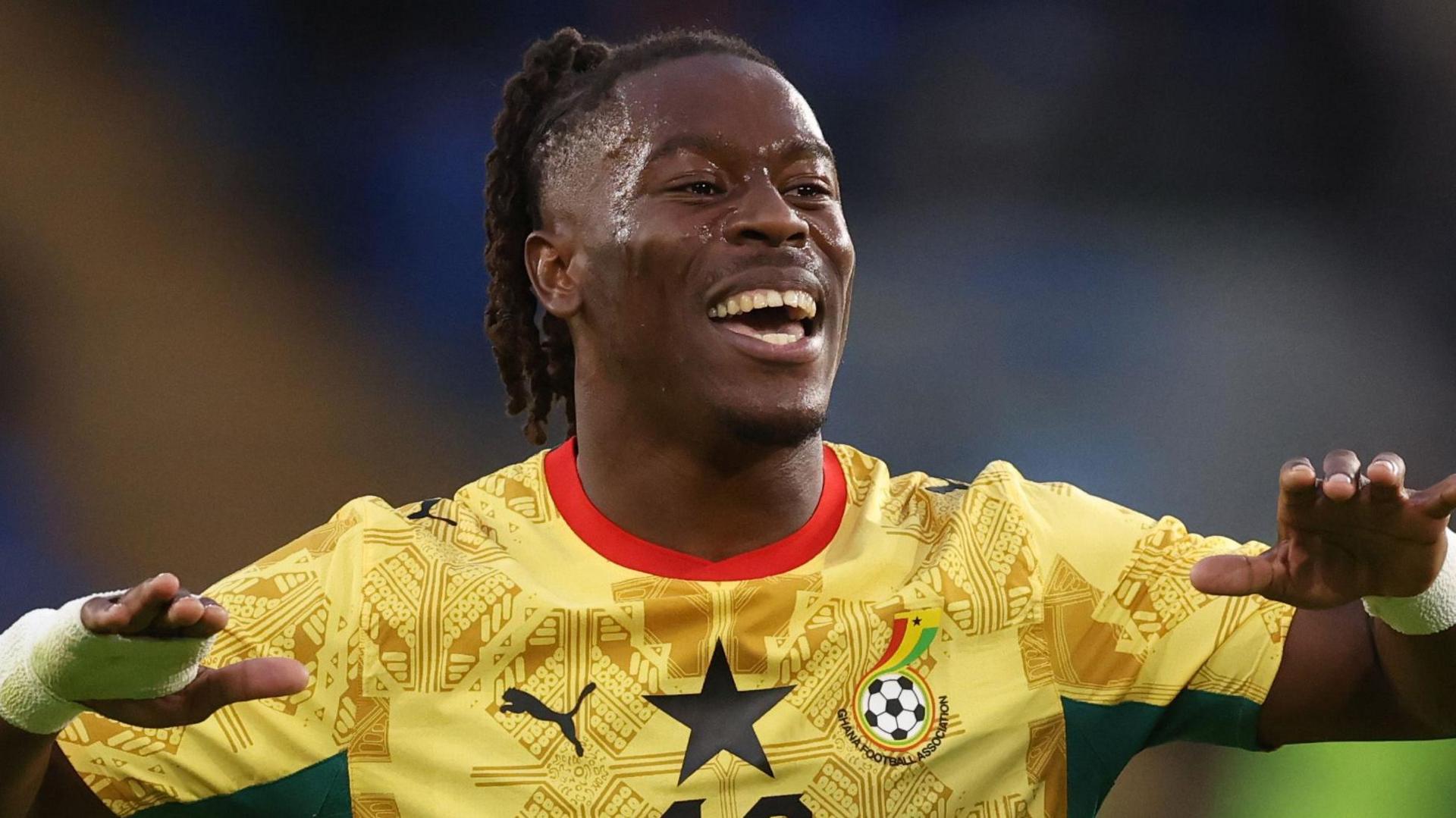 Brandon Thomas-Asante smiles and holds his hands out to his side while wearing Ghana's yellow shirt with a red trim on the neck and patterns on the main shirt.
