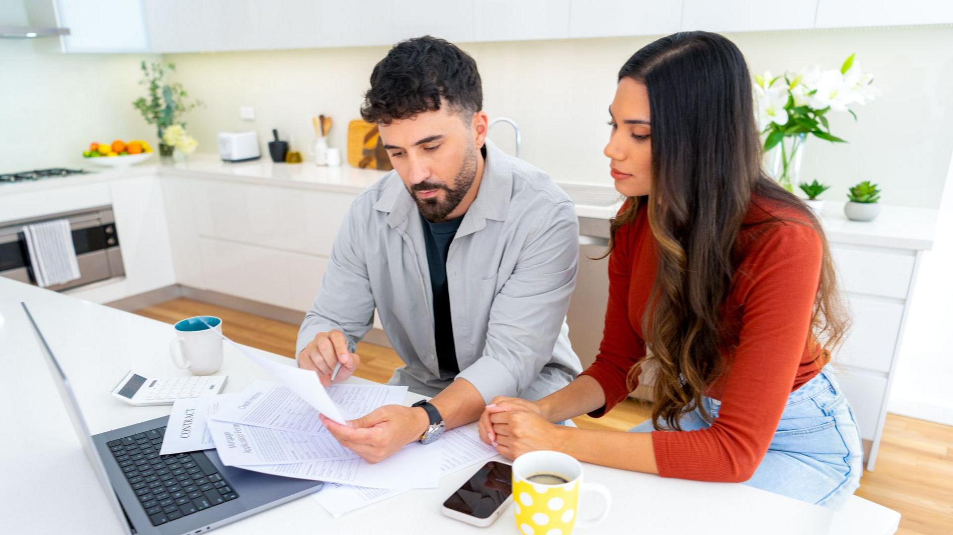 A young couple sit at their kitchen table with bills, their laptop and a calculator