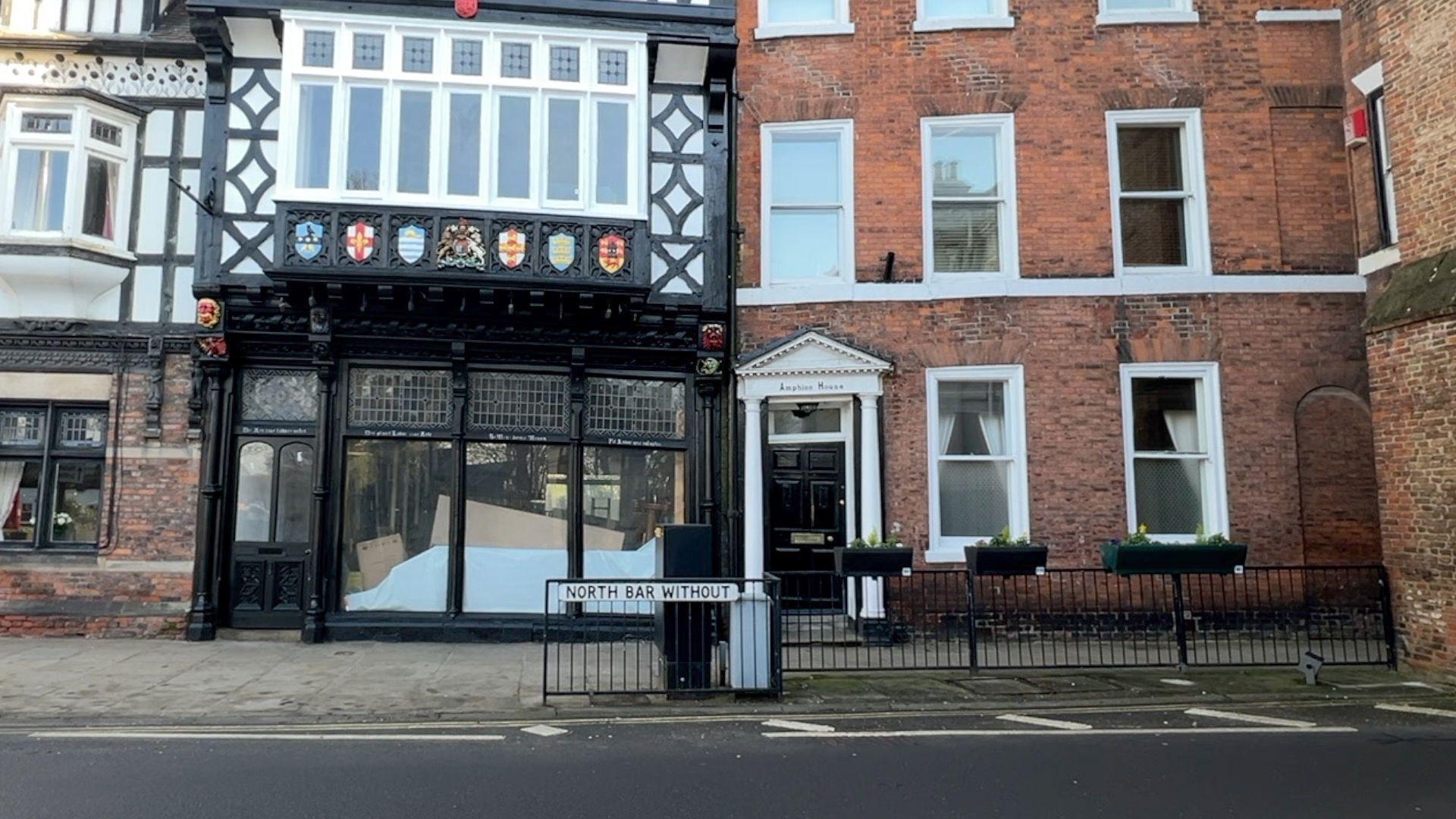 A street view of two old buildings. On the left is a tudor building in which is an empty shop. On the right is a three-storey redbrick residential property.