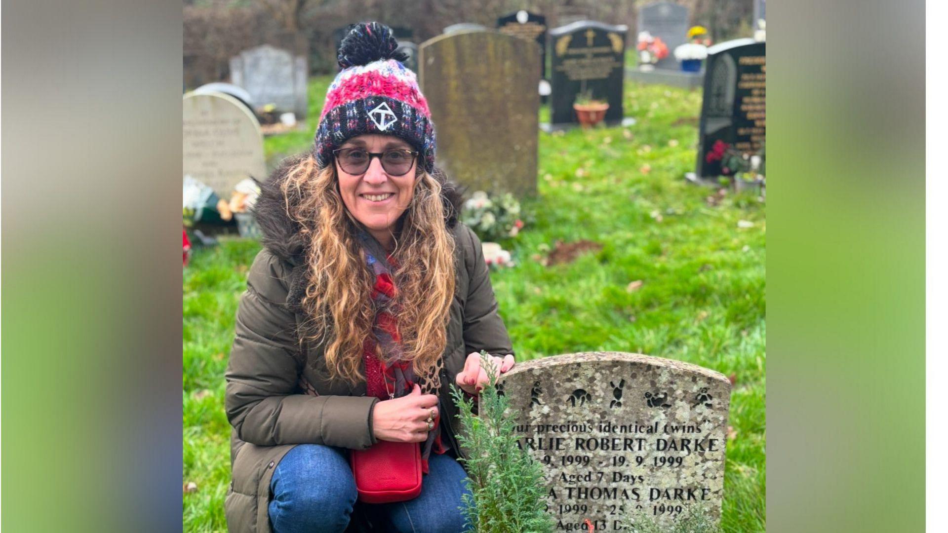 Sharon Darke with long curly hair, wearing a purple and pink woolly hat and a green coat and kneeling beside a gravestone which has the names of Charlie Robert Darke and Joshua Thomas Darke on it. There are other graves behind her