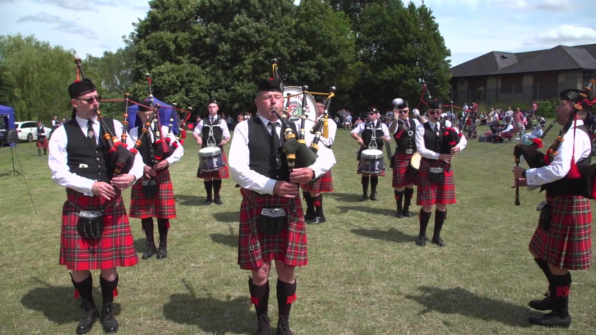 Members of a pipe band in Corby, playing the bagpipes and drums on a grass playing field. They are dressed in traditional Scottish clothing including tartan kilts.