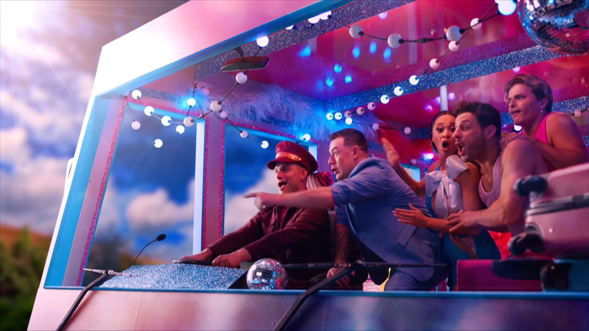 A group of dancers look surprised and point out of the front of a bus, smiling.