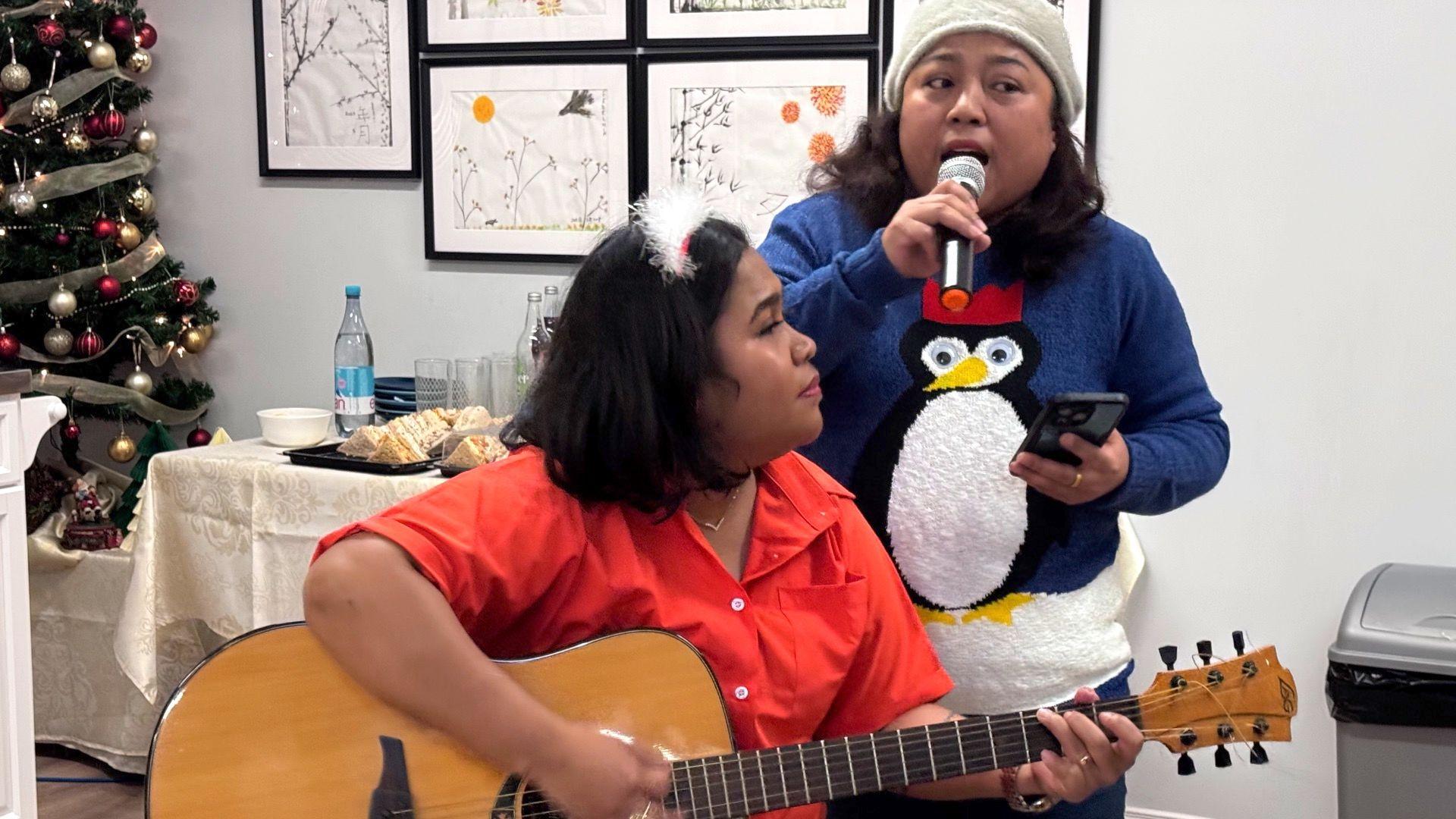 Two women - one is wearing a red blouse and is playing an acoustic guitar. The other is wearing a white woolly hat and a blue jumper with a cartoon penguin on it. She is singing into a microphone which she holds in her right hand while in her left hand she holds an iPhone. Both women have brown hair.