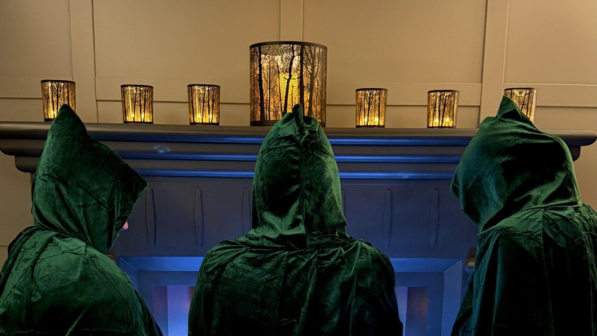The people are dressed in green velvet hoods facing towards a grey fireplace. On top of the fireplace are seven gold lanterns with the outlines of trees