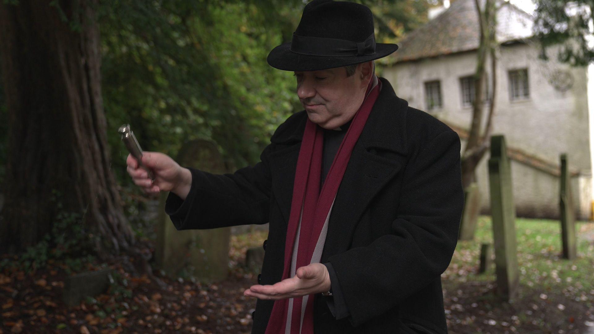 A priest wearing a trilby and long, black overcoat demonstrates a holy water sprinkler - a silver vial containing water that has been blessed.