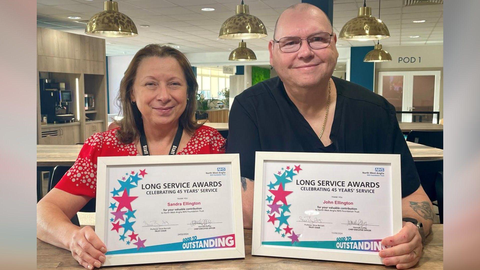 Sandy Ellington, wearing a red blouse with a white dot pattern, and John Ellington, wearing a short-sleeved black top and gold necklace are pictured with a hospital cafeteria. Both are holding their white long-service certificates and smiling.