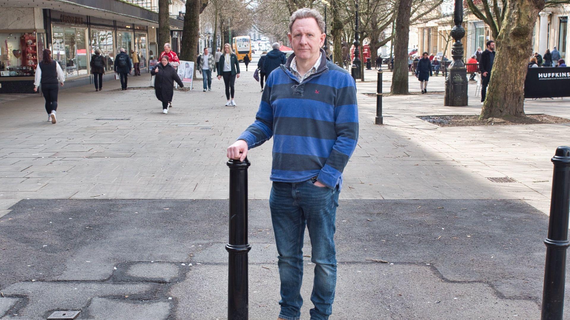 Andrew Booton standing in the middle of the promenade, wearing dark blue jeans and a blue striped jumper. He has short grey curly hair and is looking at the camera with a serious expression. His right hand is resting on top of a black bollard, while his other hand is in his front jeans pocket. Behind him you can see shoppers walking down the road. Beneath his feet there is a large patch of tarmac with has been laid to replace the broken paving stones with cover the rest of the street.