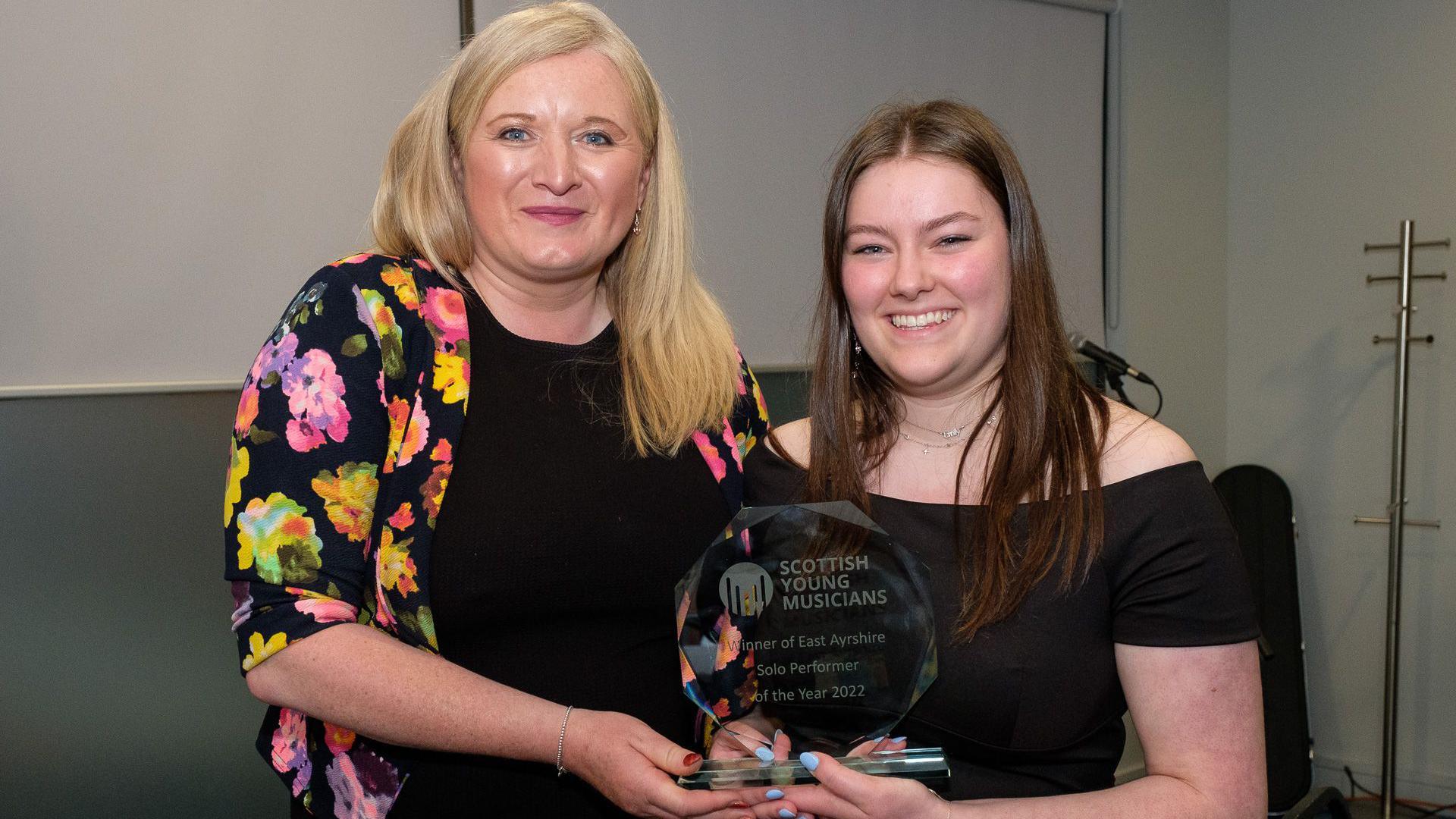 A woman with blond hair and a bright multi-coloured top on, smiling at the camera as she hands an award to a school pupil with long brown hair. Both are looking into the camera and smiling.