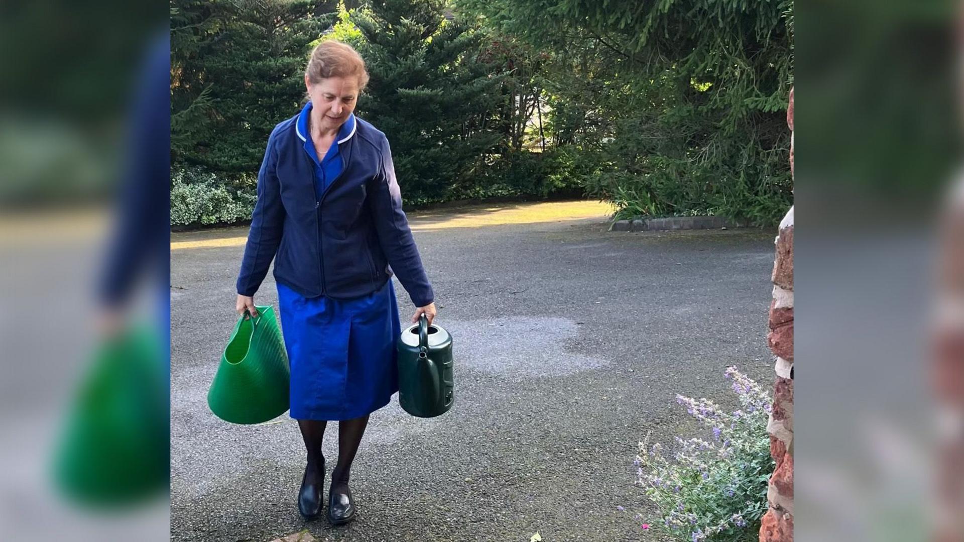 A woman walking up a driveway, carrying a trug of water and a watering can. She's wearing a royal blue nurse's uniform with a dark blue coat over the top. 