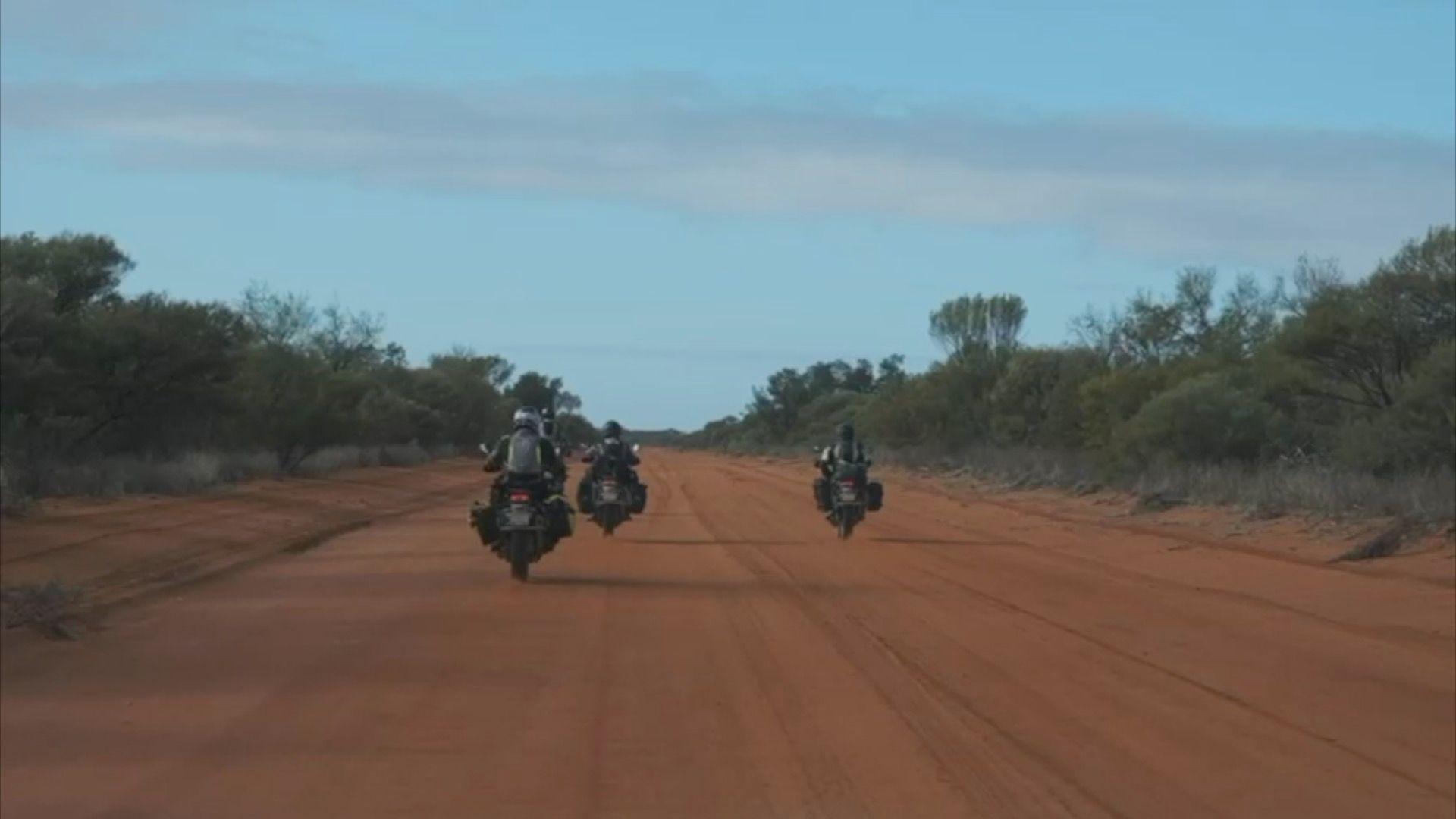 Three motorcycle riders riding along a large sandy path, surrounded by trees