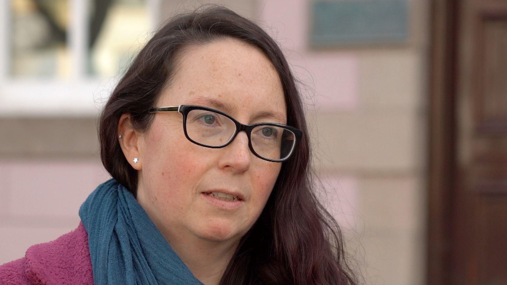 Deputy Louise Doublet is wearing a pink coat, a blue scarf and glasses. She has brown hair 