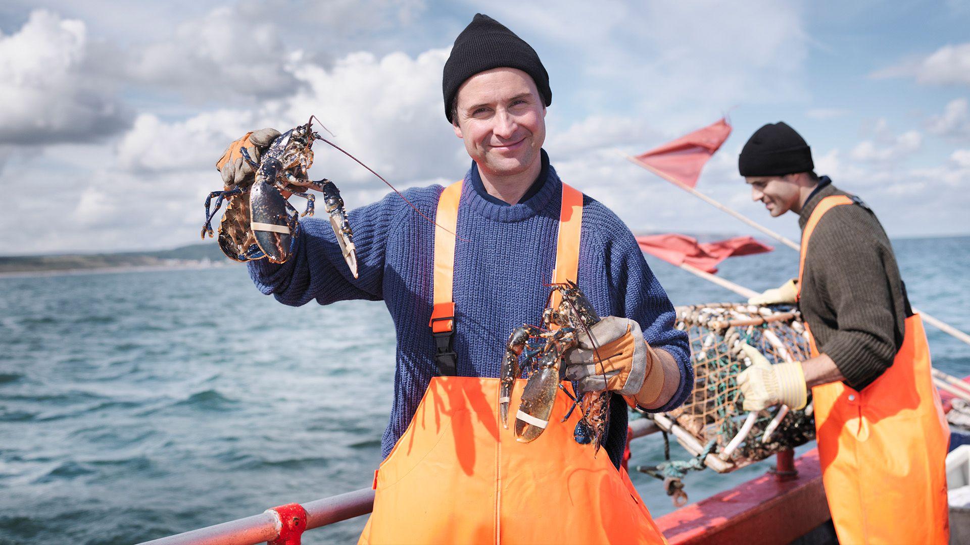 A fisherman holds up two lobsters while at sea on a boat. He is wearing a blue knitted jumper and bright orange overalls