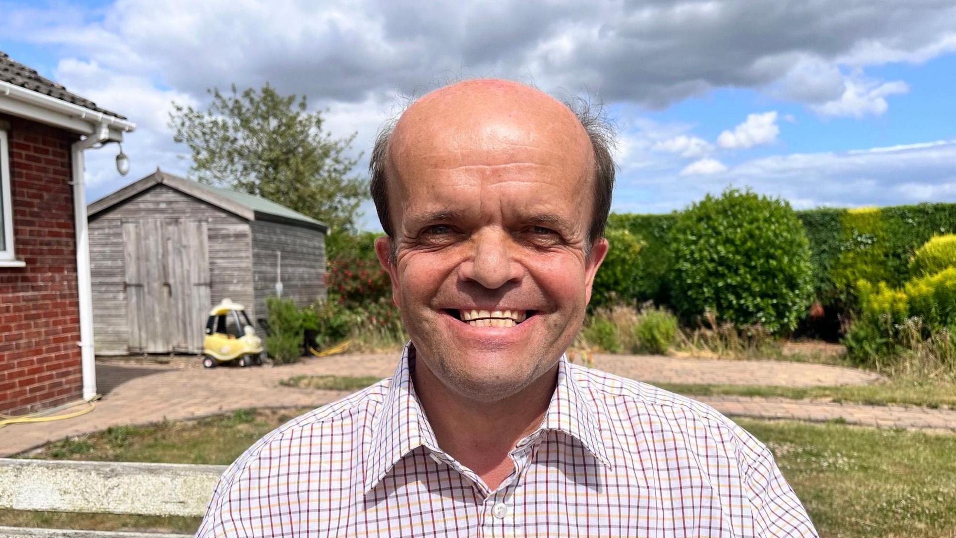 A bald smiling man in a tattersall checked shirt is standing in a garden, there's a wooden shed behind him and a child's toy car parked in front of it.