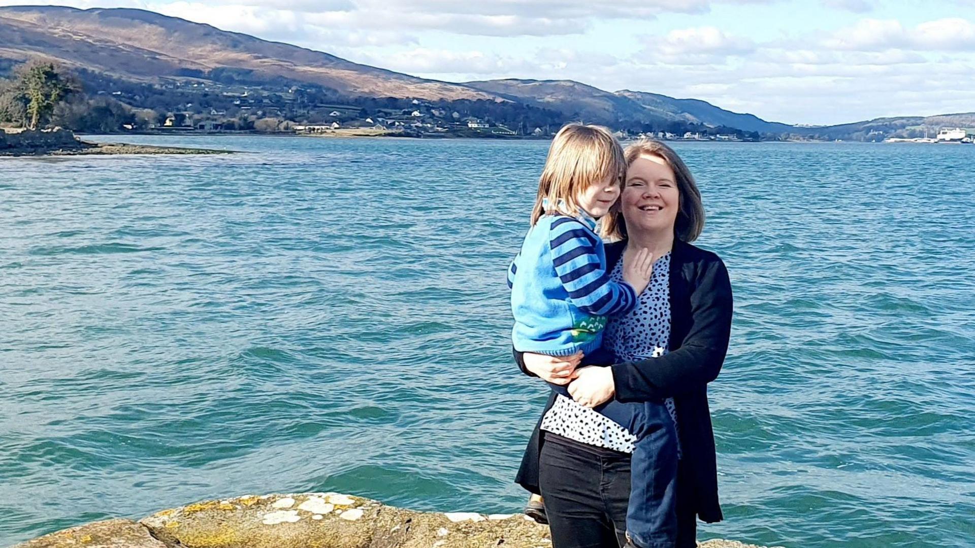 Fay and her young son Charlie at a shoreline on a sunny day. Fay has shoulder-length fair hair and is wearing a black cardigan, black jeans and a blue spotted top. She is carrying Charlie in her arms. He has long fair hair covering his eyes. He is wearing a blue and navy jumper with striped arms and dark blue trousers.