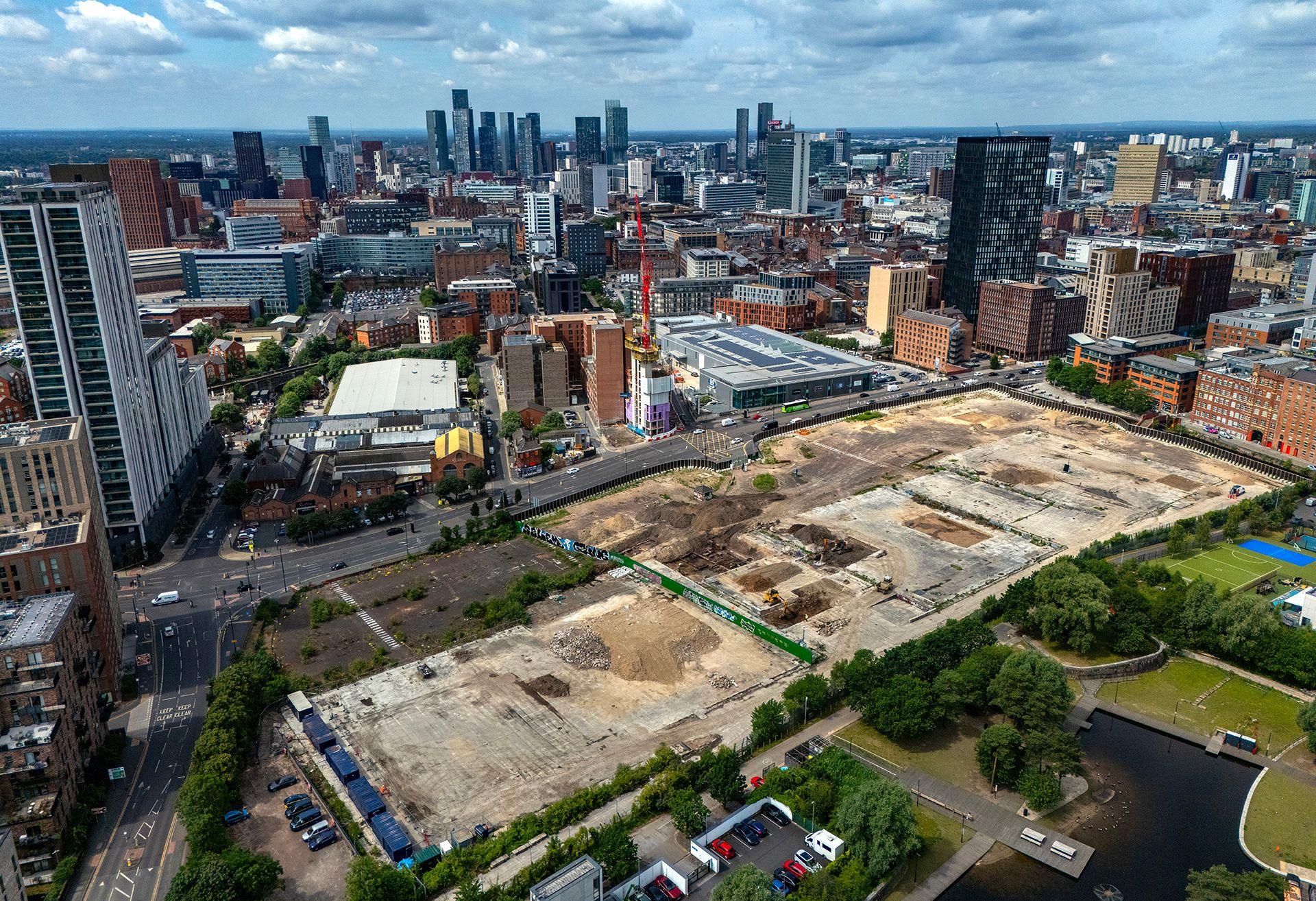 An aerial photograph taken with a drone shows construction on a large, brownfield site in Manchester. Tower blocks can be seen in the distance, smaller office blocks are nearby and there is a multi-lane road running along the left hand side. A red crane stands in the middle of the picture.