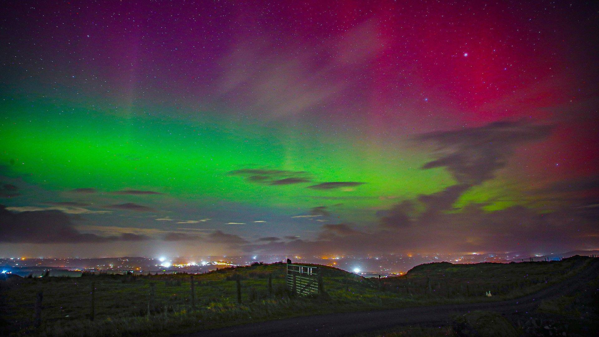 A landscape photo taken at night from a hilled area showing the Northern Lights in the sky. There are streaks of green, red and purple lights interrupted by some clouds.