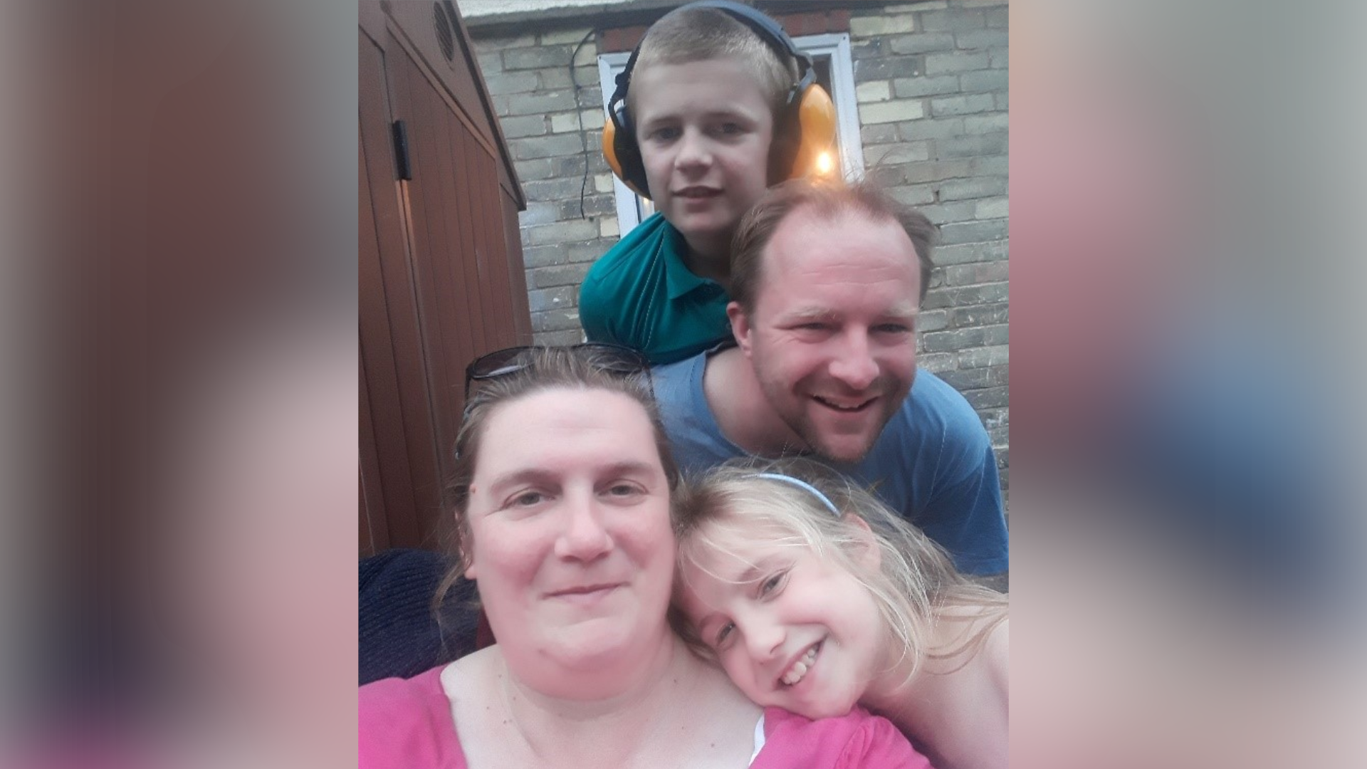 Marianne with her family. They have taken a selfie-style photo and are all smiling at the camera. Marianne has blonde hair and is resting her head on her mother's shoulder who has dark hair and is wearing a pink top. Marianne's father stands above them in a blue top while her brother Levi stands next to him wearing protective headphones for his ears.