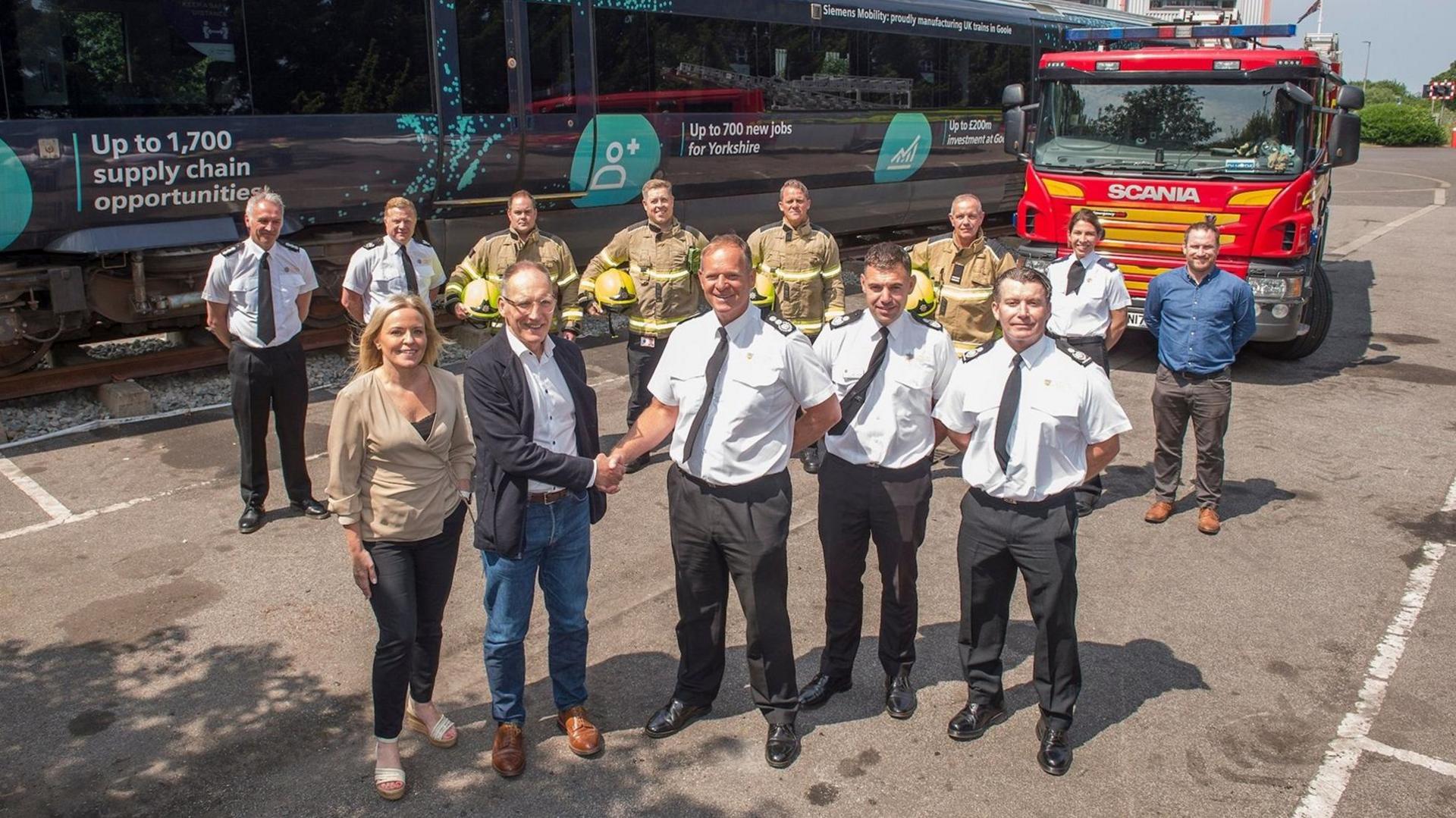 Firefighters, some wearing hi-vis workwear, with staff from Siemens Mobility in front of a train and a fire engine. A senior fire officer is shaking hands with a man from Siemens Mobility.