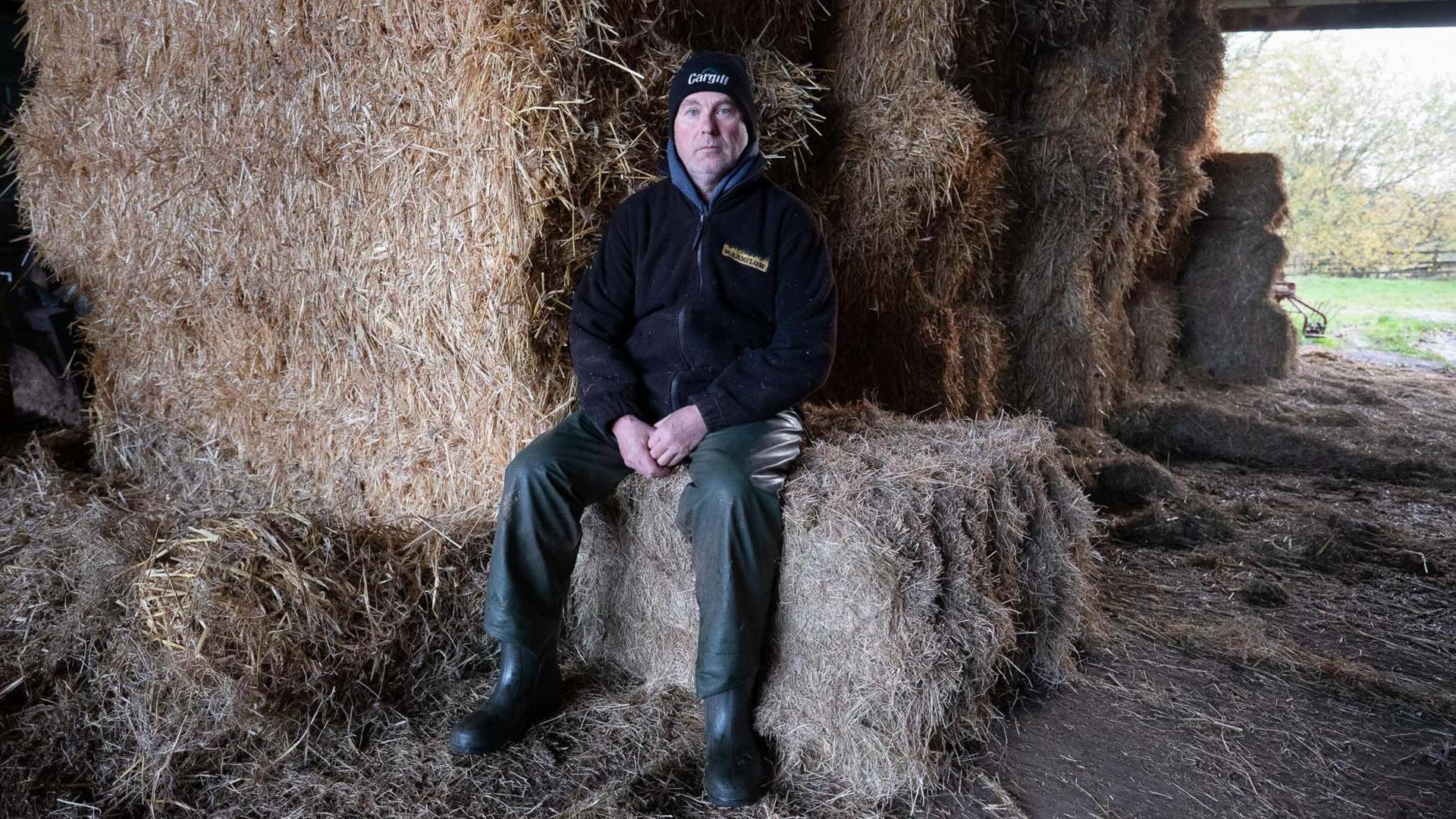 Andrew Jackson sitting on a hay bale, with other hay bales around him. He is looking at the camera with a blank face. He is wearing a woolly hat, fleece and wellies.