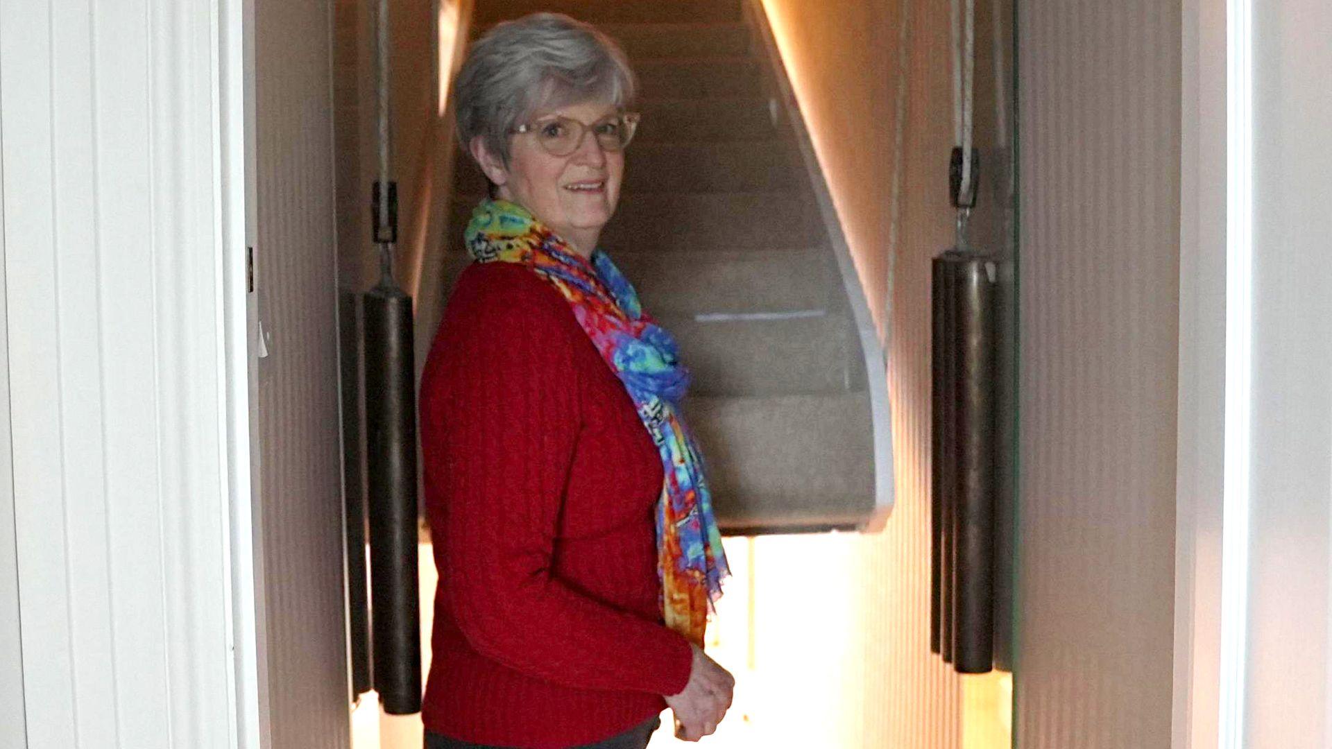 Judith standing by her counter-weight staircase. The staircase is partly risen and has a cream carpet on it, with two weights to the left and right. She is wearing a red jumper, multi-coloured scarf and dark trousers. 