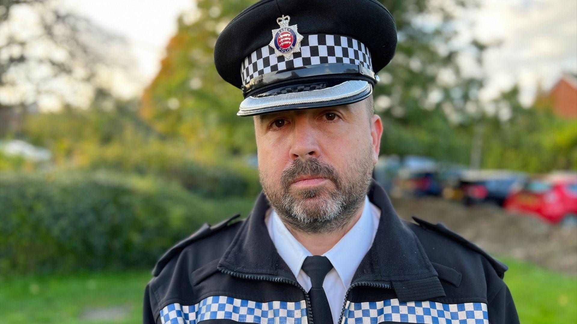 Ch Supt Stuart Hooper from Essex Police. He is wearing  a police uniform, which includes a black peaked cap, a black jacket with a line of light blue and white check, a white shirt and black tie. He is standing in front of greenery.