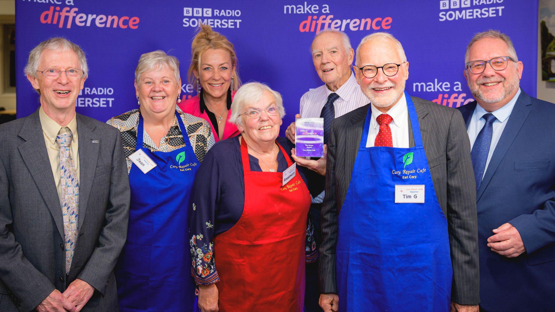 A mix of six elderly men and women wearing aprons look at camera in front of TV presenter Sarah Beeny. Behind them is a promotion wall with "make a difference" and "BBC Somerset" written in pale text on a blue background in multiple places