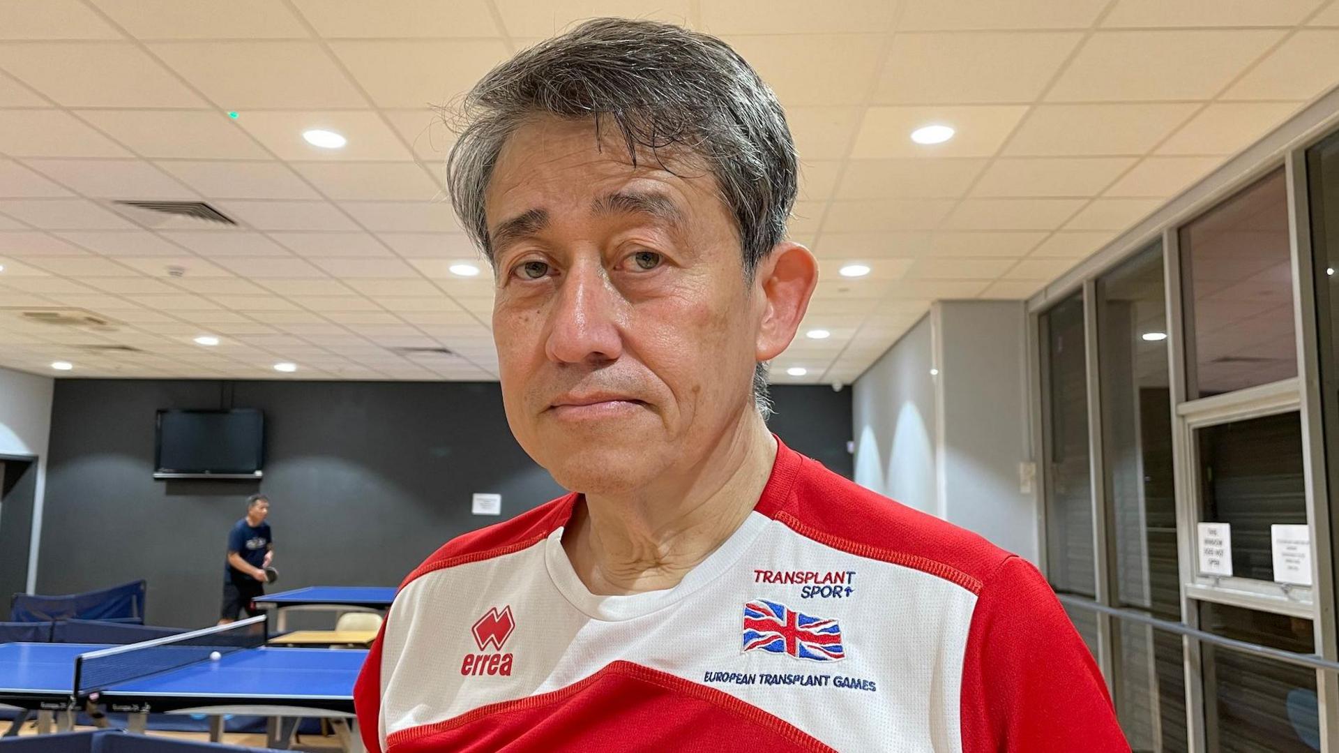 Alex Bell has short black-grey hair and brown eyes and is looking directly into the camera. He's wearing a red-and-white t-shirt bearing the Union Jack, a 'TRANSPLANT SPORT' logo and the words 'EUROPEAN TRANSPLANT GAMES'. In the background there are table tennis tables and a man in a blue T-shirt holding a table tennis paddle.