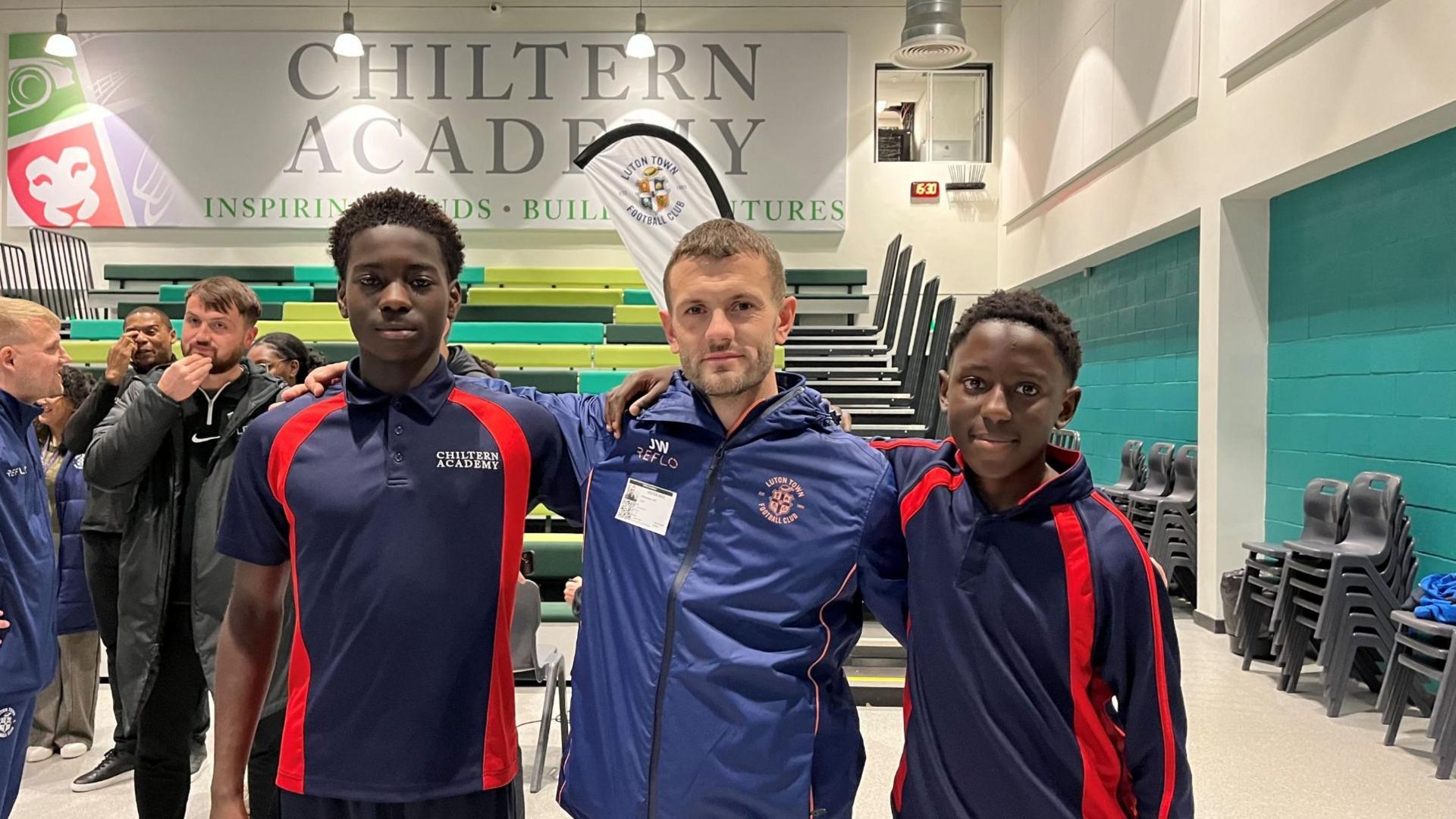 Luton Football Club's manager Jack Wilshere, in the middle appearing with short crew-cut hair and wearing a blue Luton Town-branded jacket, poses with two male pupils from Chiltern Academy in a sports hall. The two children are wearing navy and red Chiltern Academy branded clothing.