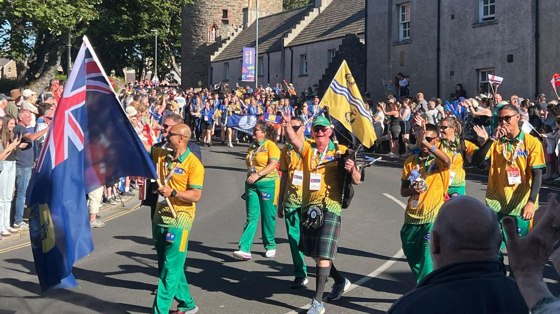 Athletes walk down the middle of the road waving flags in tracksuits. Crowds cheer at the side of the road.