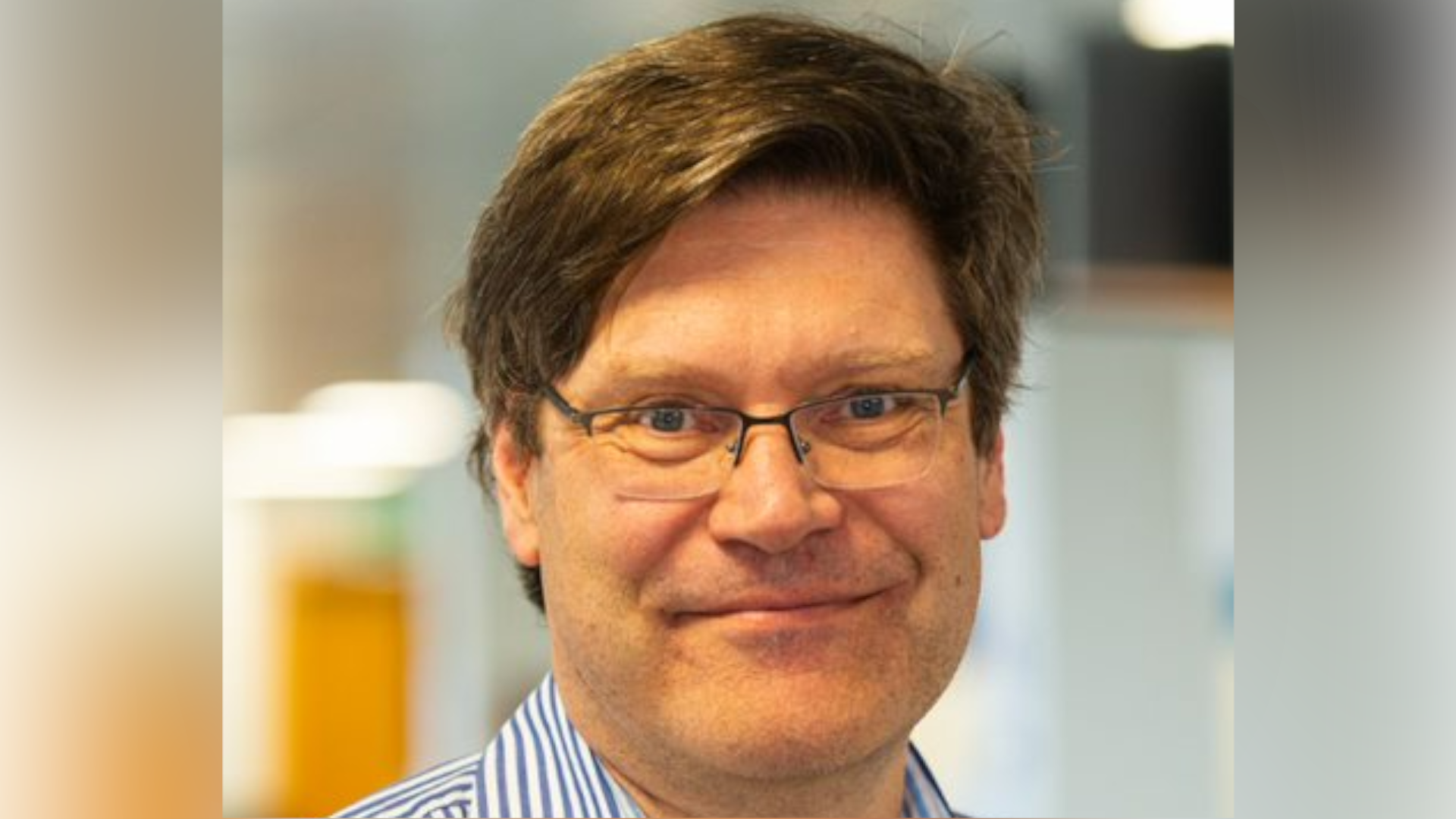 James Allen smiles at the camera. He has brown hair and wears glasses as well as a blue and white striped shirt.