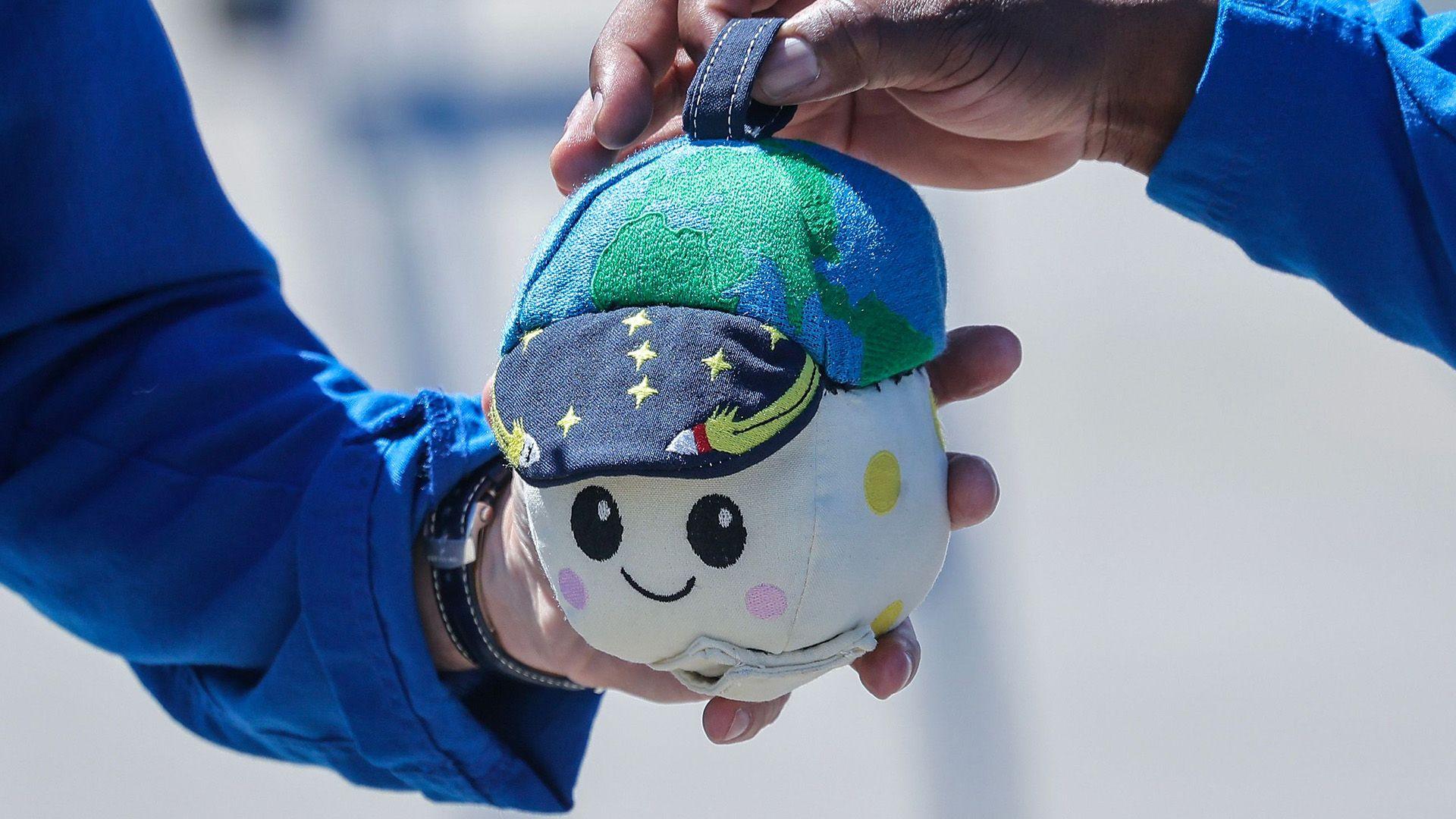 A plushie is being held by two astronauts. Its is a round beige moon with a smiley face and a hat that looks like the Earth.