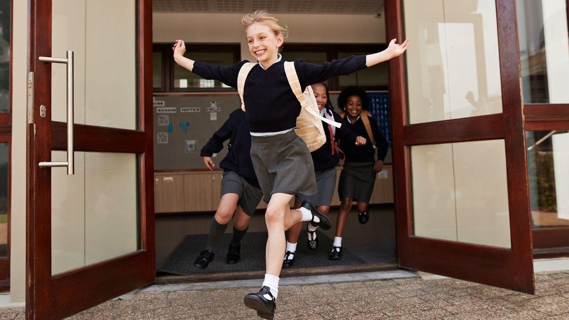 Girl runs out of the school entrance with her arms in the air and a big smile on her face. She is wearing her school uniform and other pupils are following her