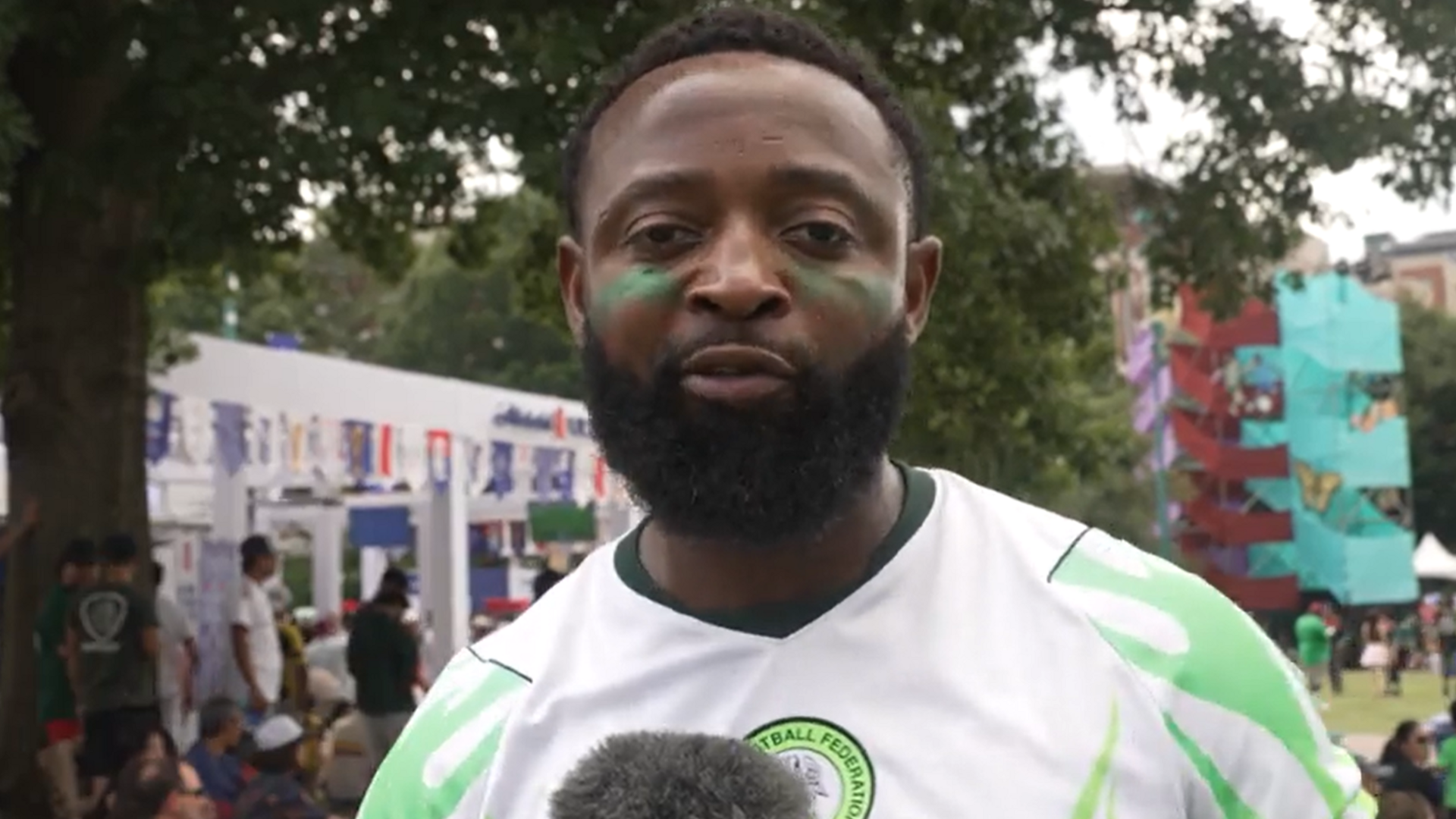 Nigeria fan Kingsley, visible from chest up wearing a white and green Nigeria shirt, looks straight towards the camera as he gives an interview. A tree and several people in a park can be seen out of focus in the background