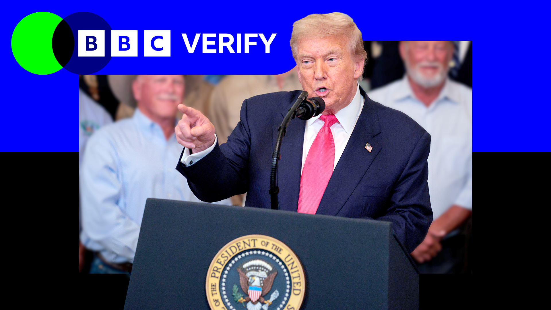 Donald Trump speaking at a lectern bearing the seal of the president of the United States. He is wearing a dark suit and red tie while a small crowd is standing behind him. 
