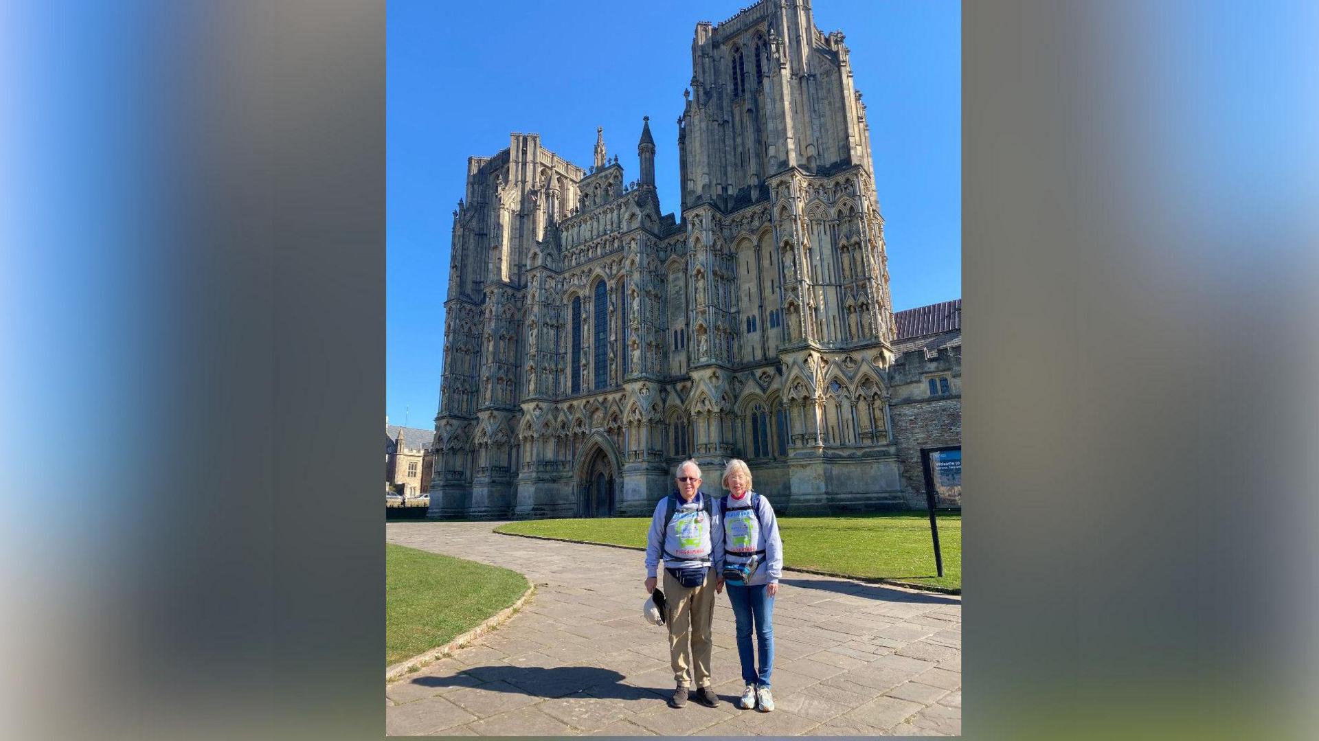 The Banhams in front of a large stone-built cathedral. It is the eastern facade, with an entrance and towers. There is a lawn and a path in front, where Mr and Mrs Banham are standing with their grey sweatshirts on.